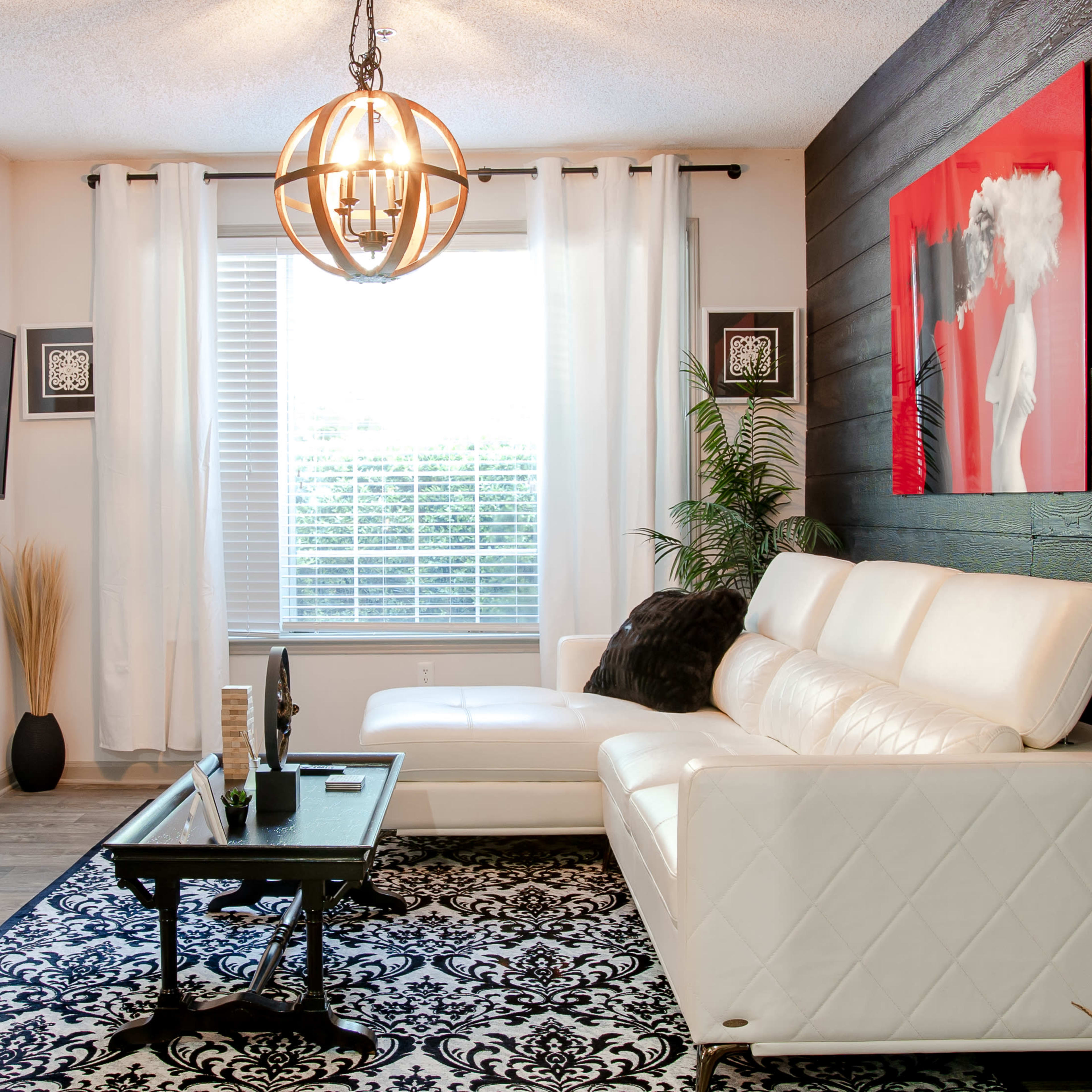 A modern living room features a white sectional sofa, a black coffee table, and a large window with sheer curtains, illuminated by a round pendant light.