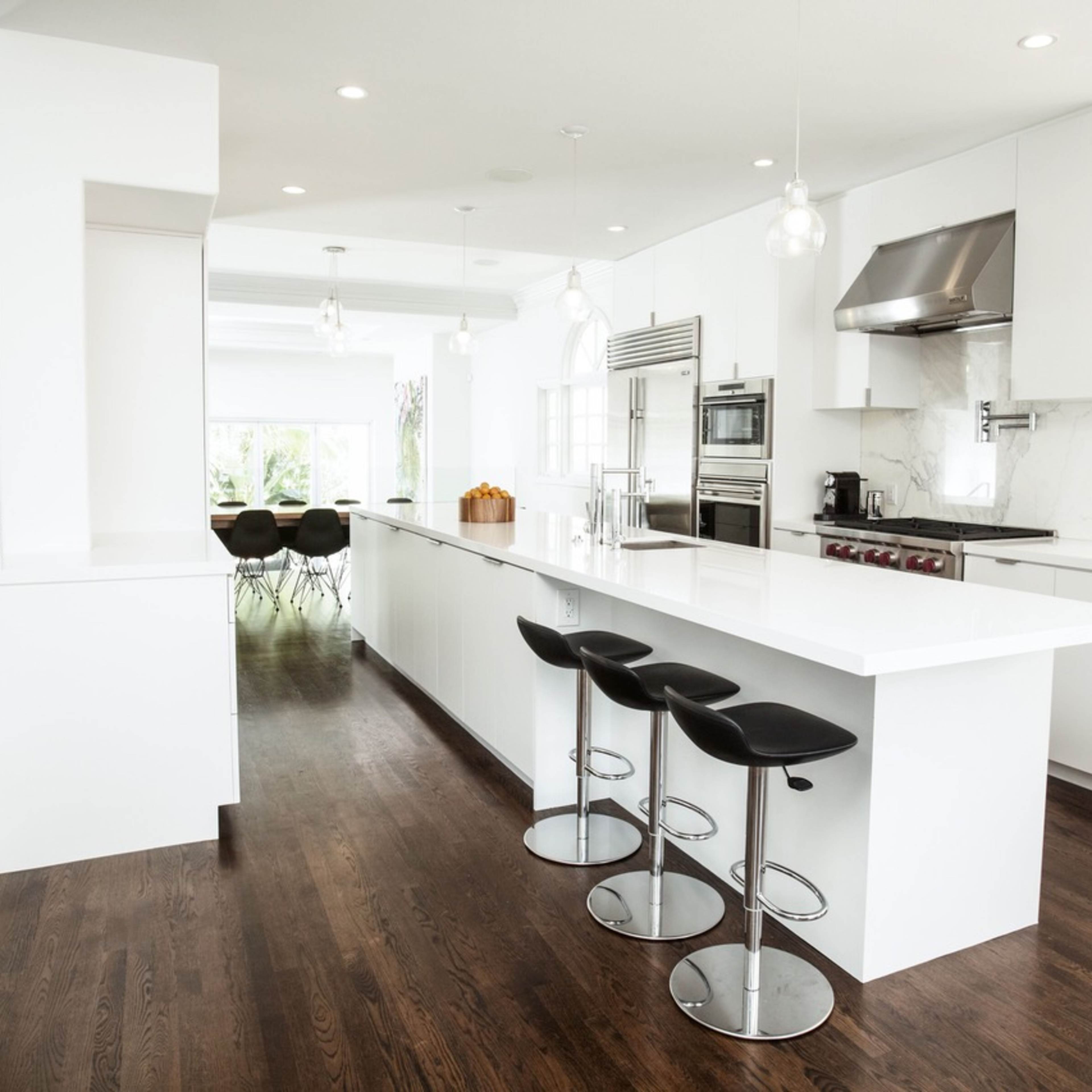 A modern kitchen features a white island counter with black bar stools, stainless steel appliances, and large windows allowing natural light to flood the space.