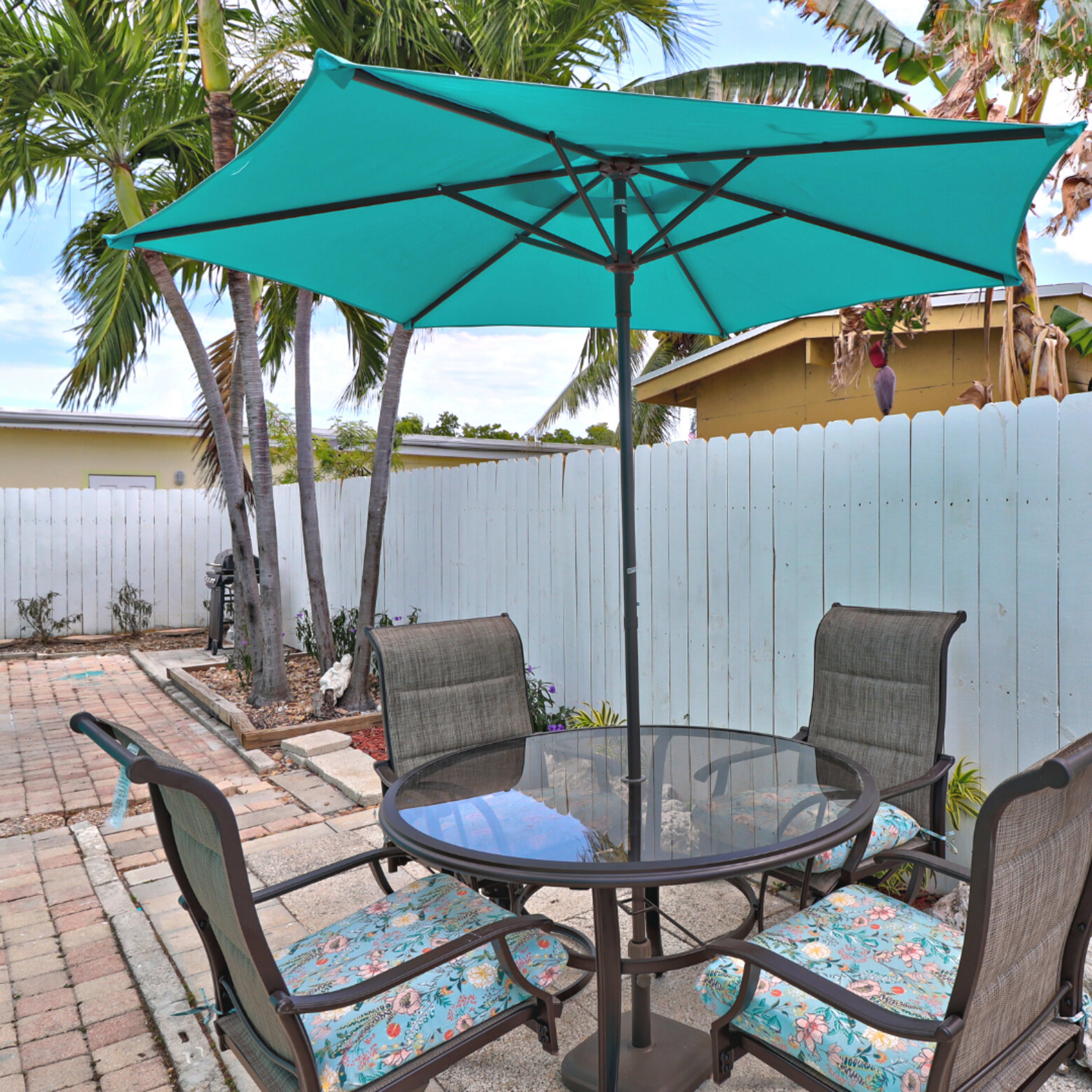 A round glass table with four chairs is shaded by a teal umbrella in a small yard surrounded by a white fence and palm trees.