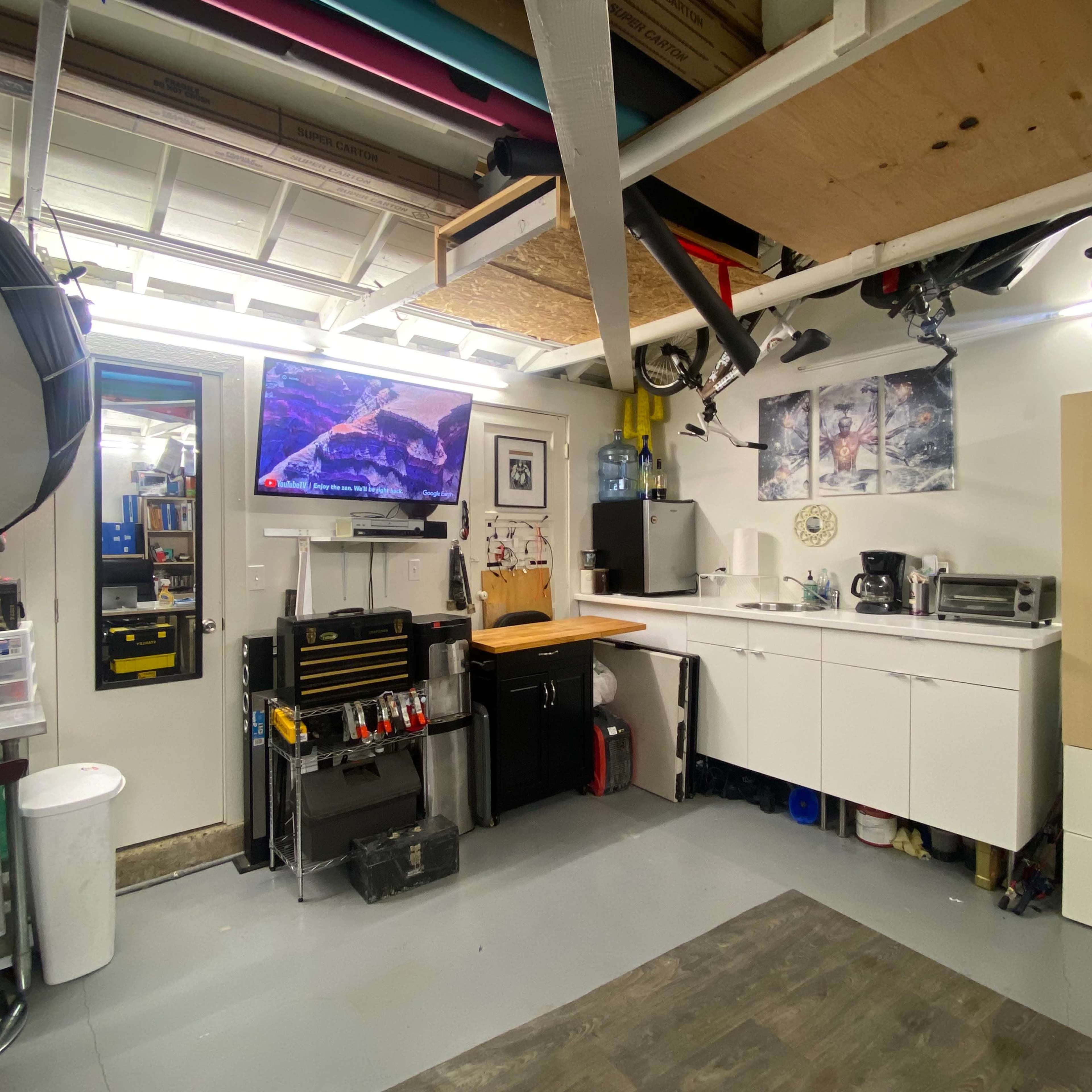 A well-organized garage or workshop space featuring a wall-mounted television, storage cabinets, workbench, and various tools.