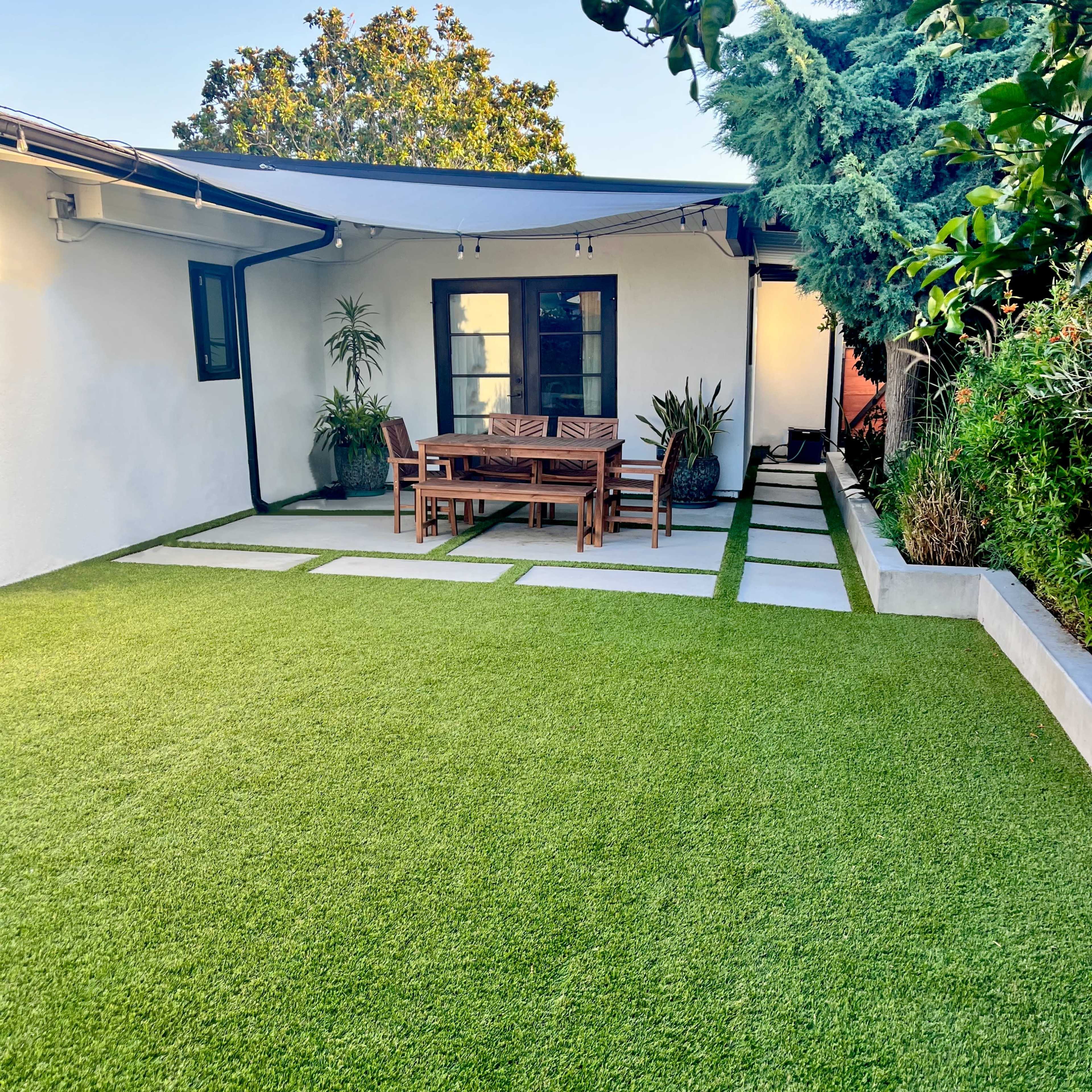 A backyard patio featuring a wooden dining table and chairs on a well-maintained artificial lawn, adjacent to a house with a modern exterior.