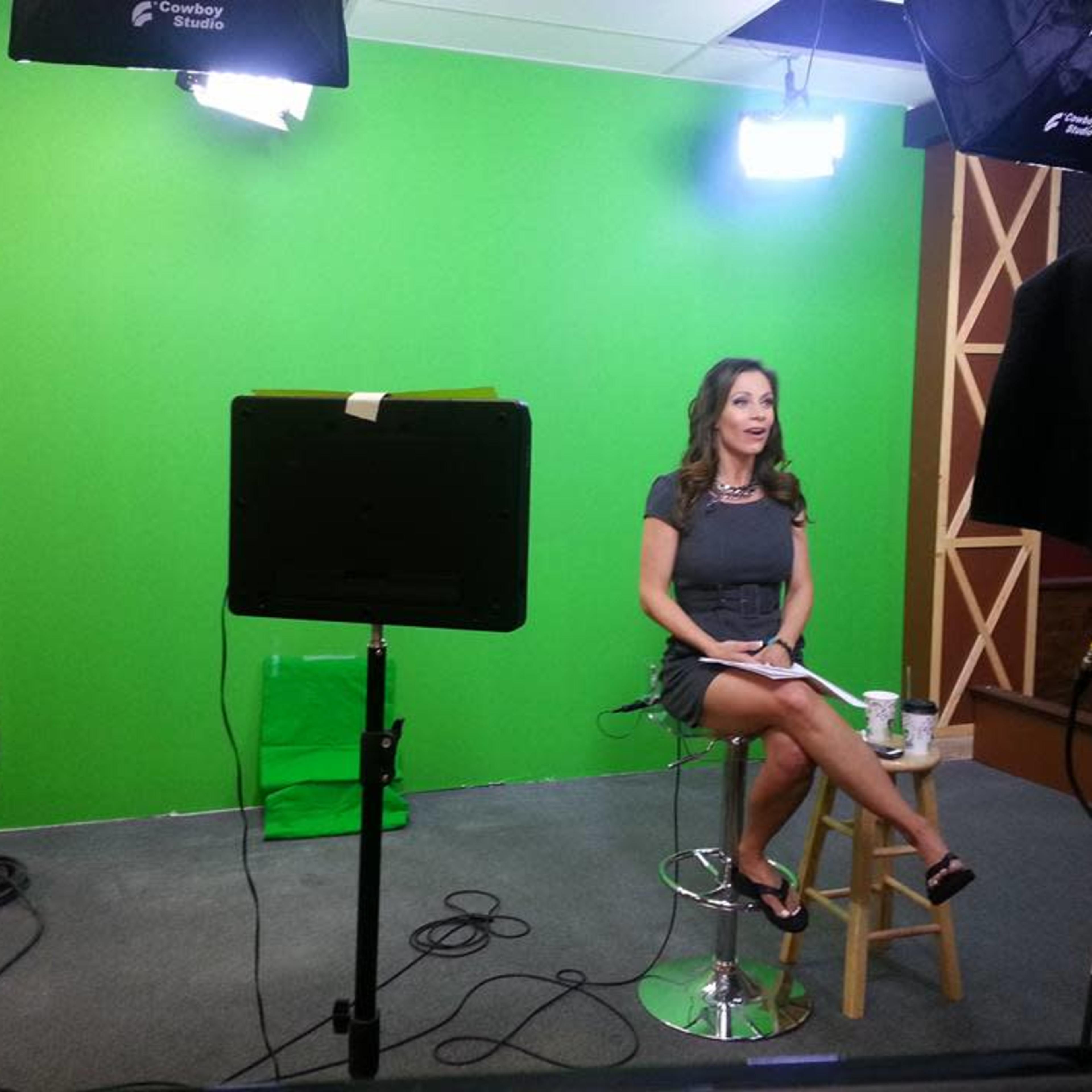 A woman sits on a stool in front of a green screen, with studio lights and cameras positioned around her.
