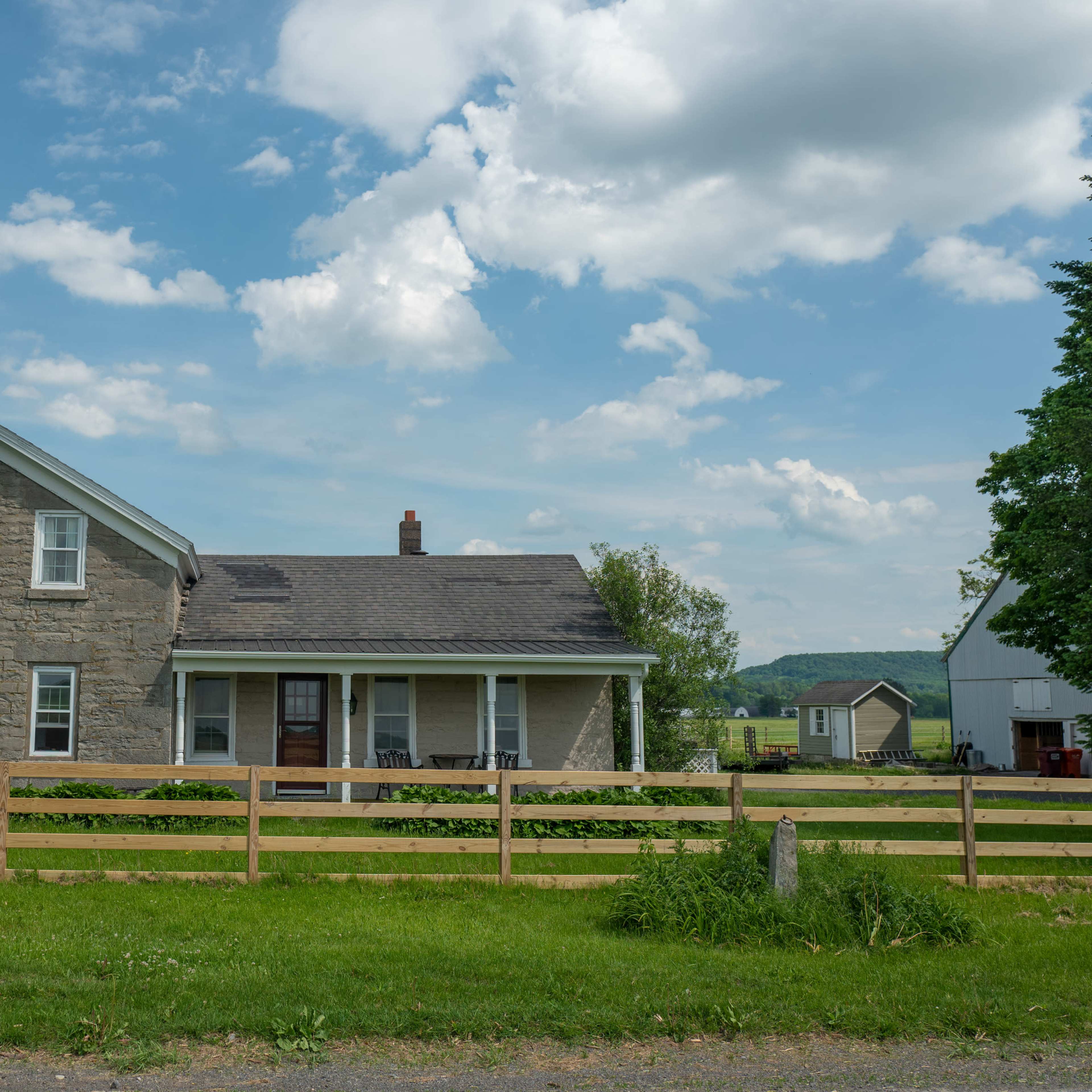 A two-story stone farmhouse with a porch is shown beside a wooden fence, set against a backdrop of green fields and cloudy skies.
