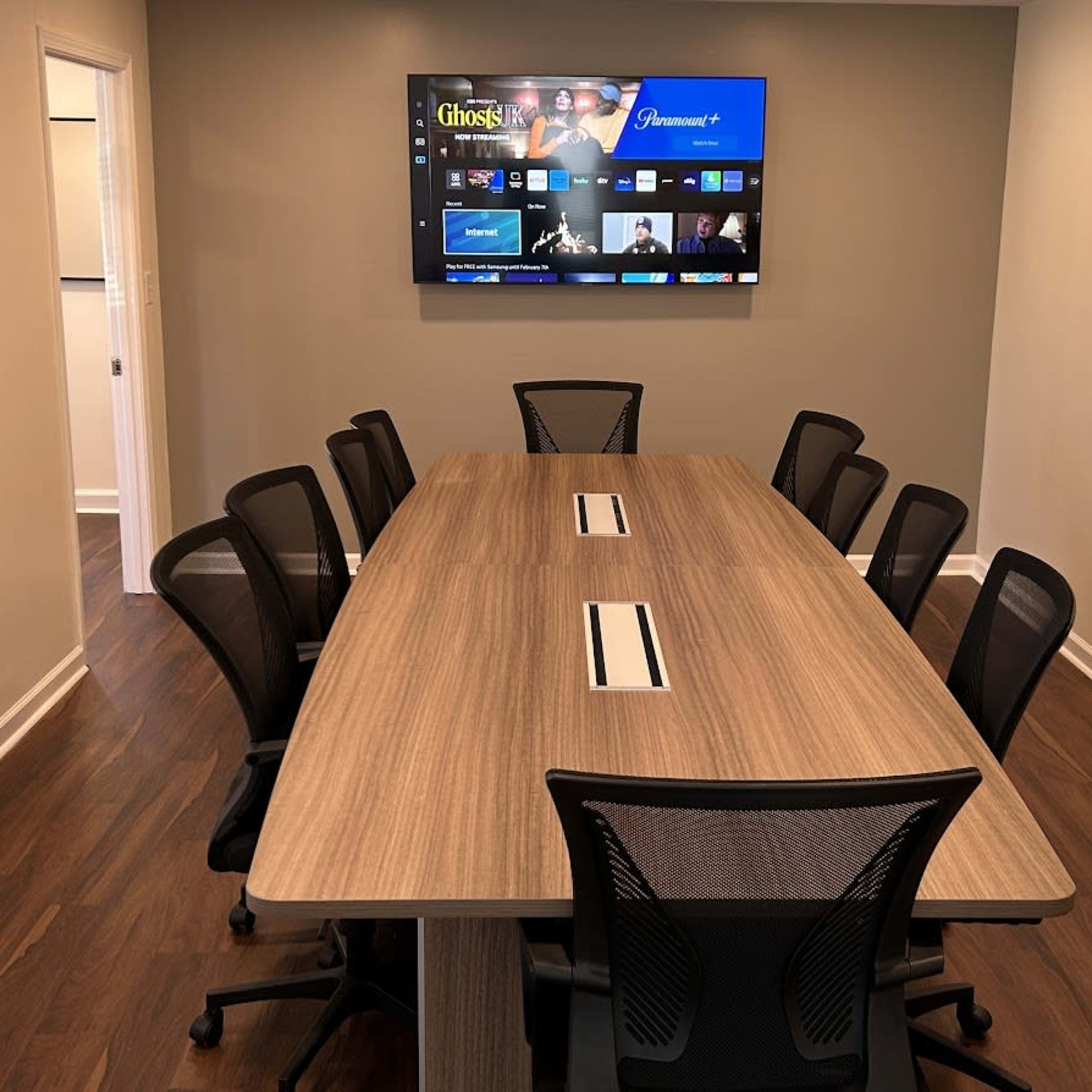 A conference room features a large wooden table surrounded by eight black office chairs, with a wall-mounted screen displaying a video call interface.