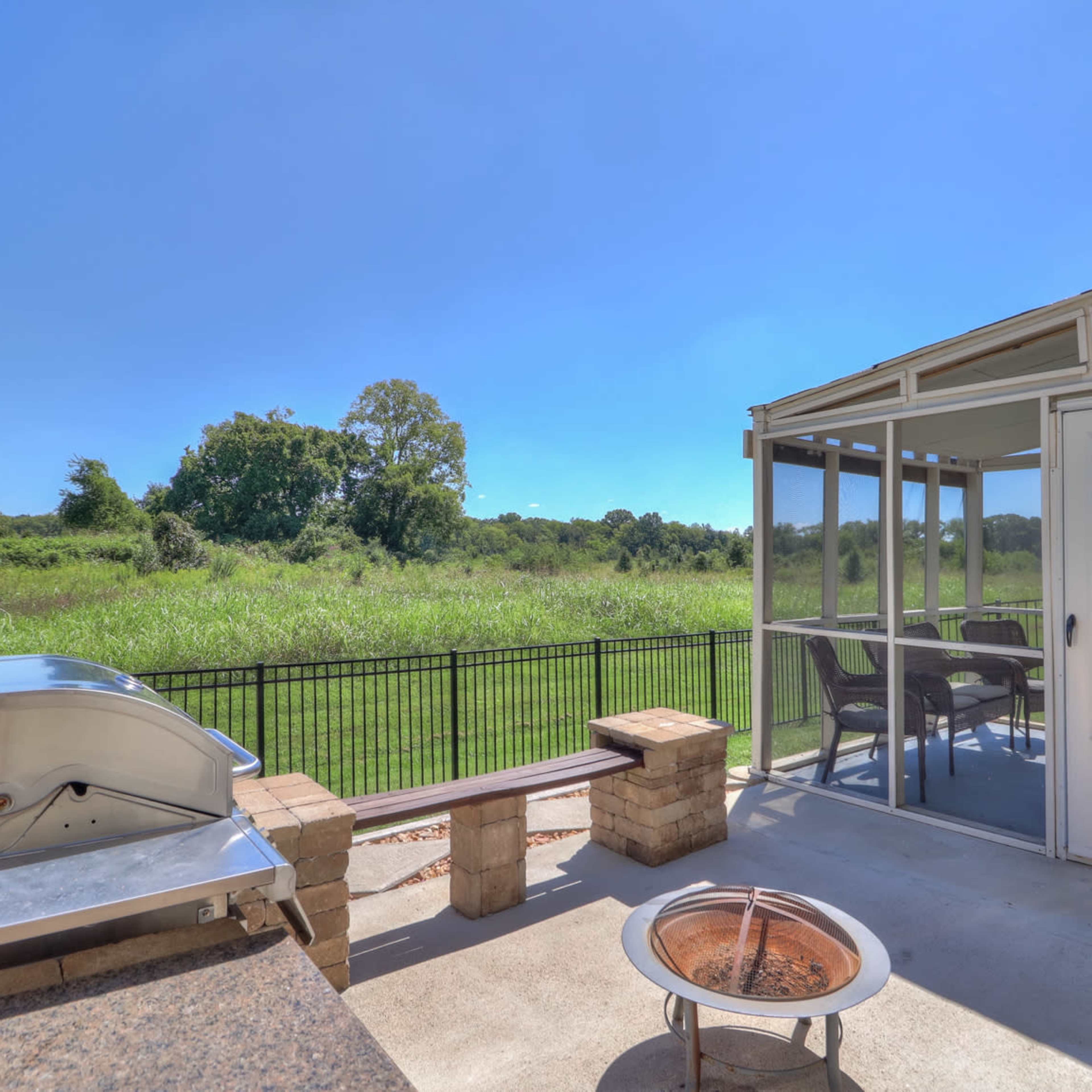 A backyard patio with a built-in grill, a fire pit, and a screened-in area, overlooking a grassy field.