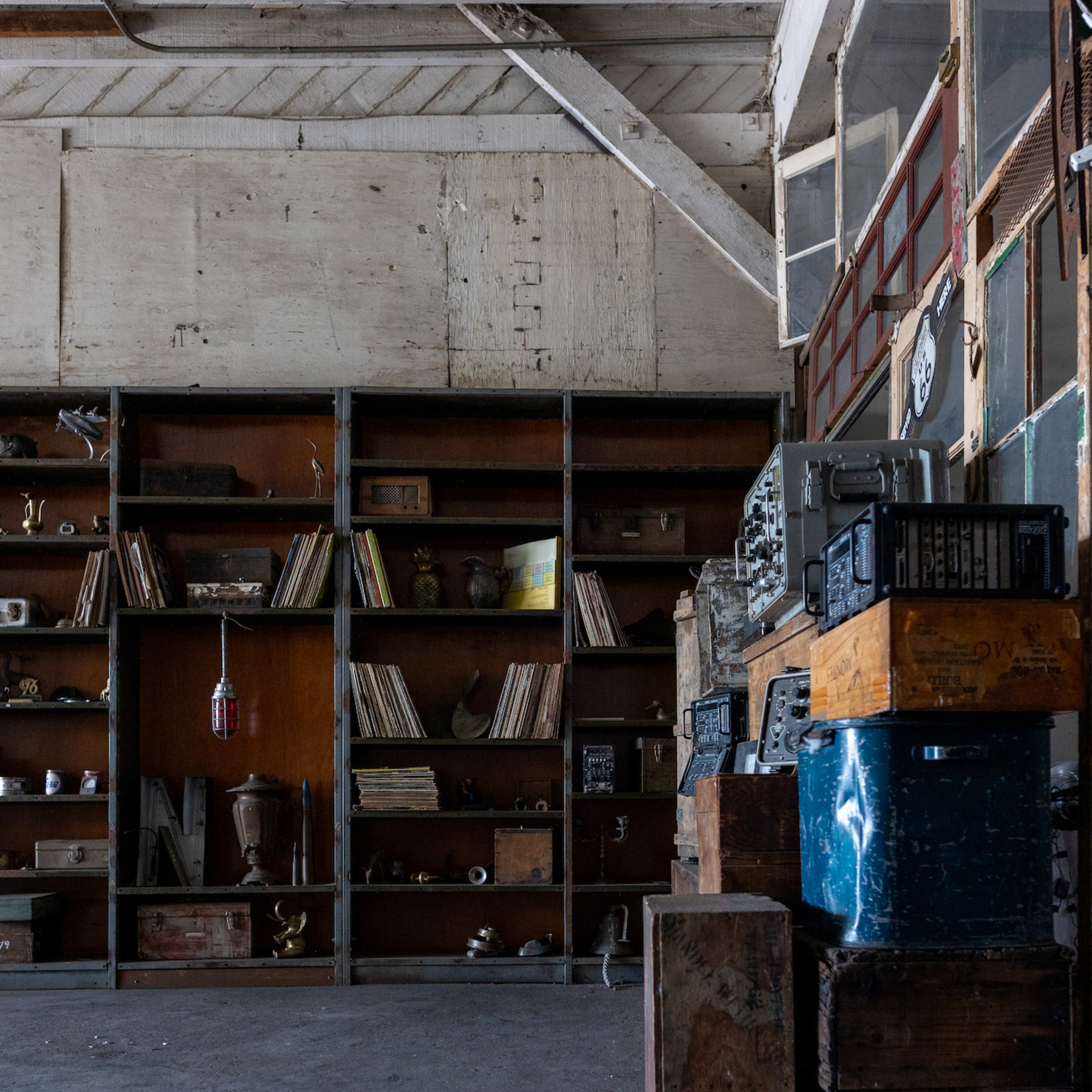 A room with a wooden floor, featuring a tall shelf filled with books and various objects, alongside stacked wooden crates.
