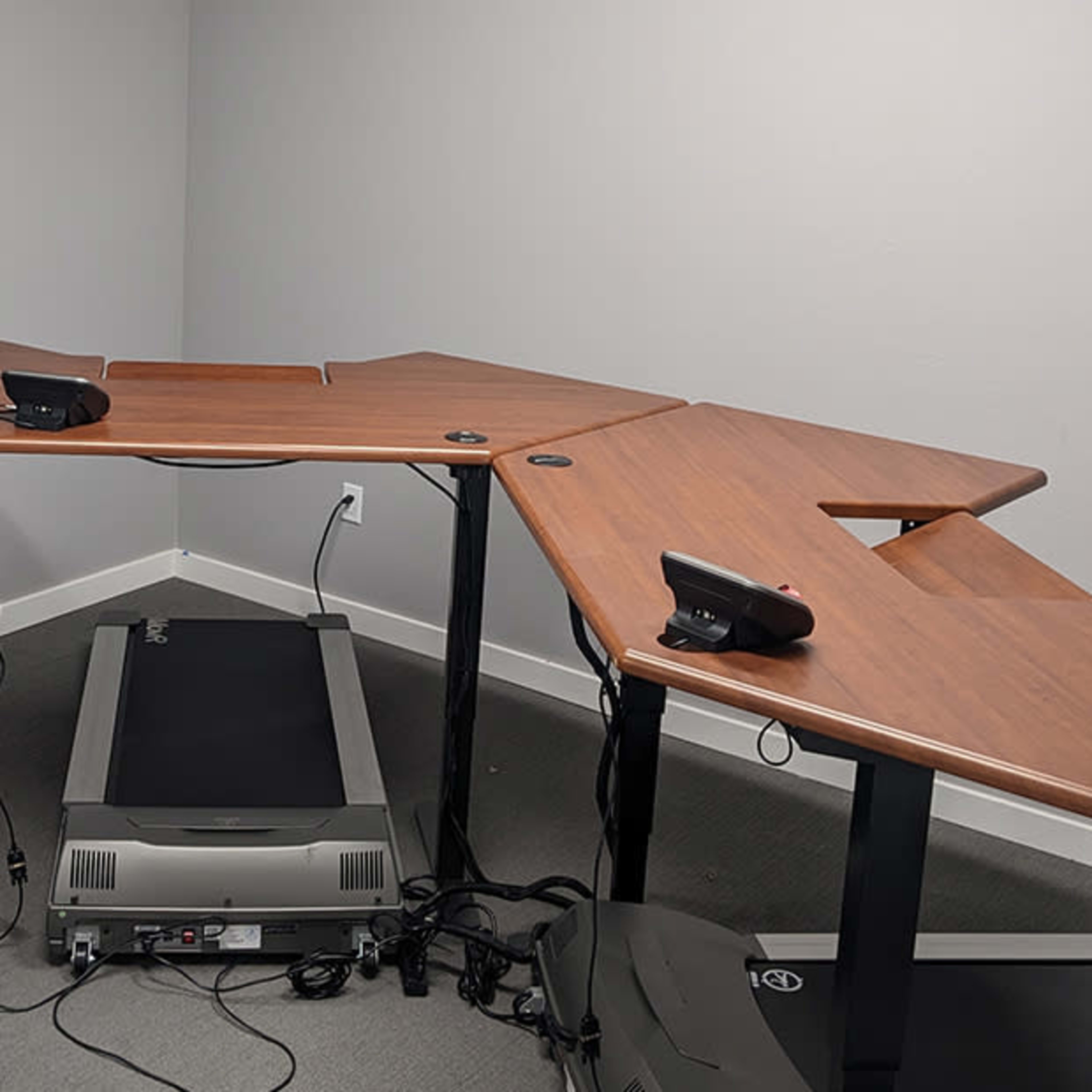 A multi-segmented wooden desk with telephone equipment sits in a corner of a room, next to a treadmill and black office chairs.