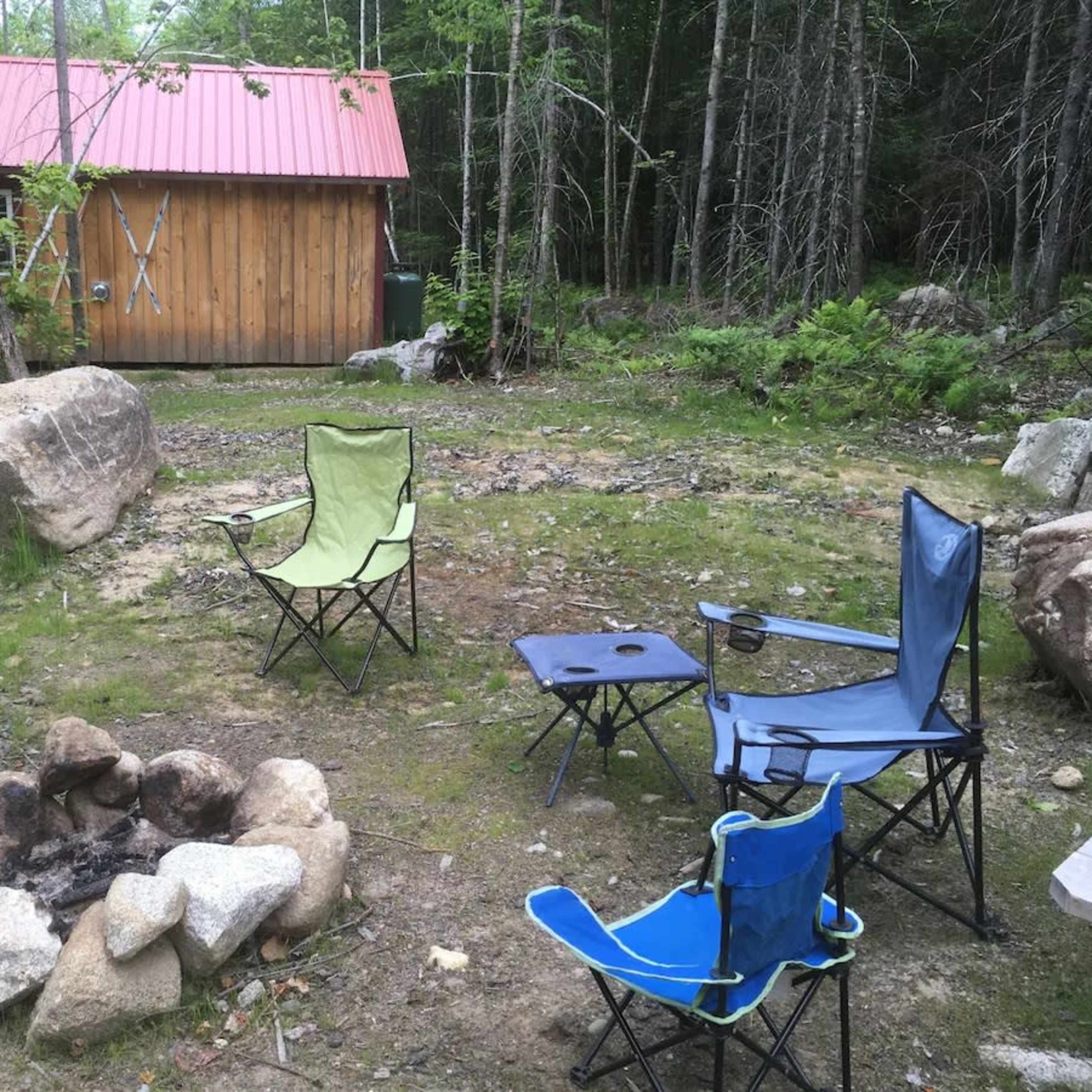A campfire area with several folding chairs set around a stone fire pit, next to a wooden shed in a wooded environment.