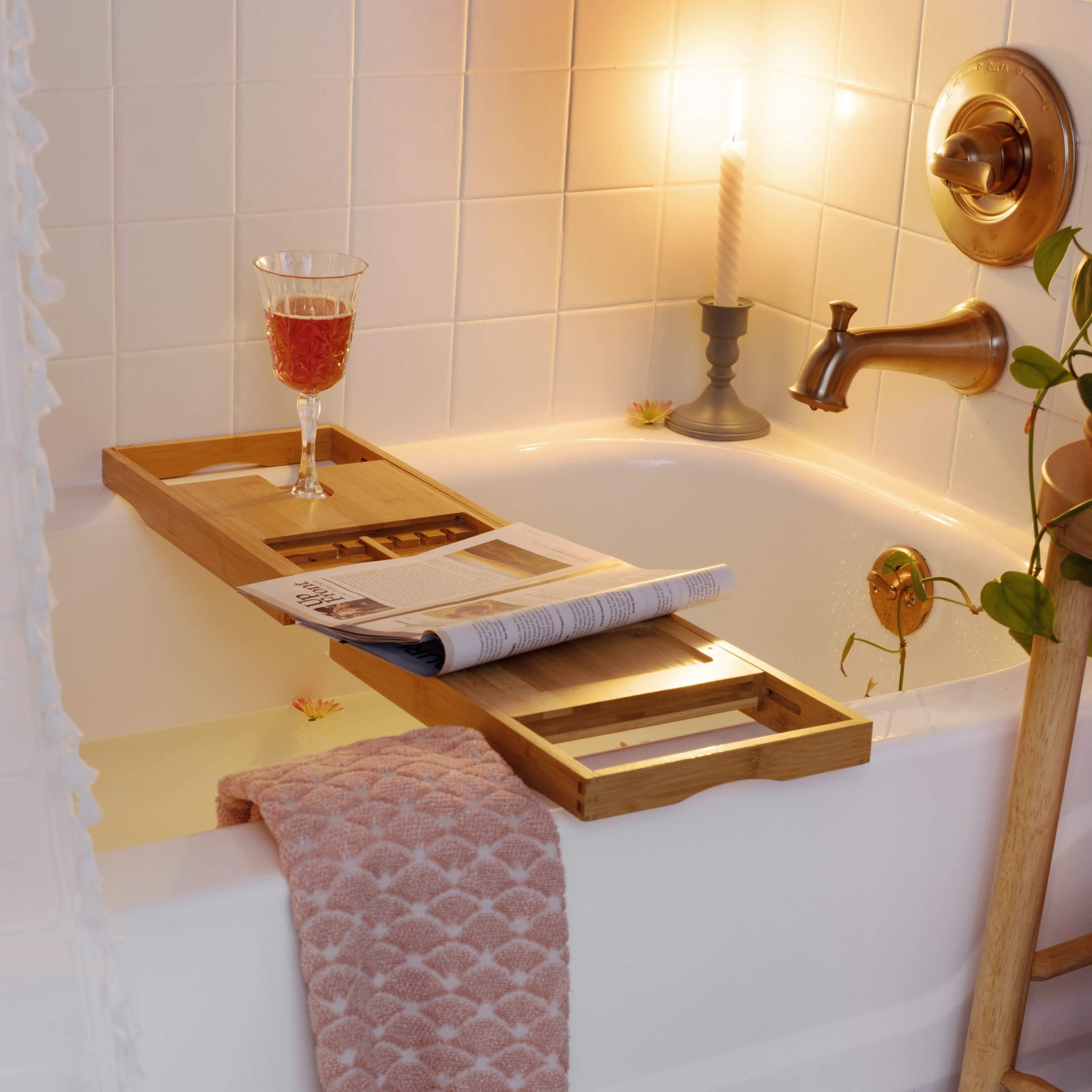 A bathtub is adorned with a wooden tray holding a glass of drink and a magazine, while a pink towel drapes over the side and a candle illuminates the scene.