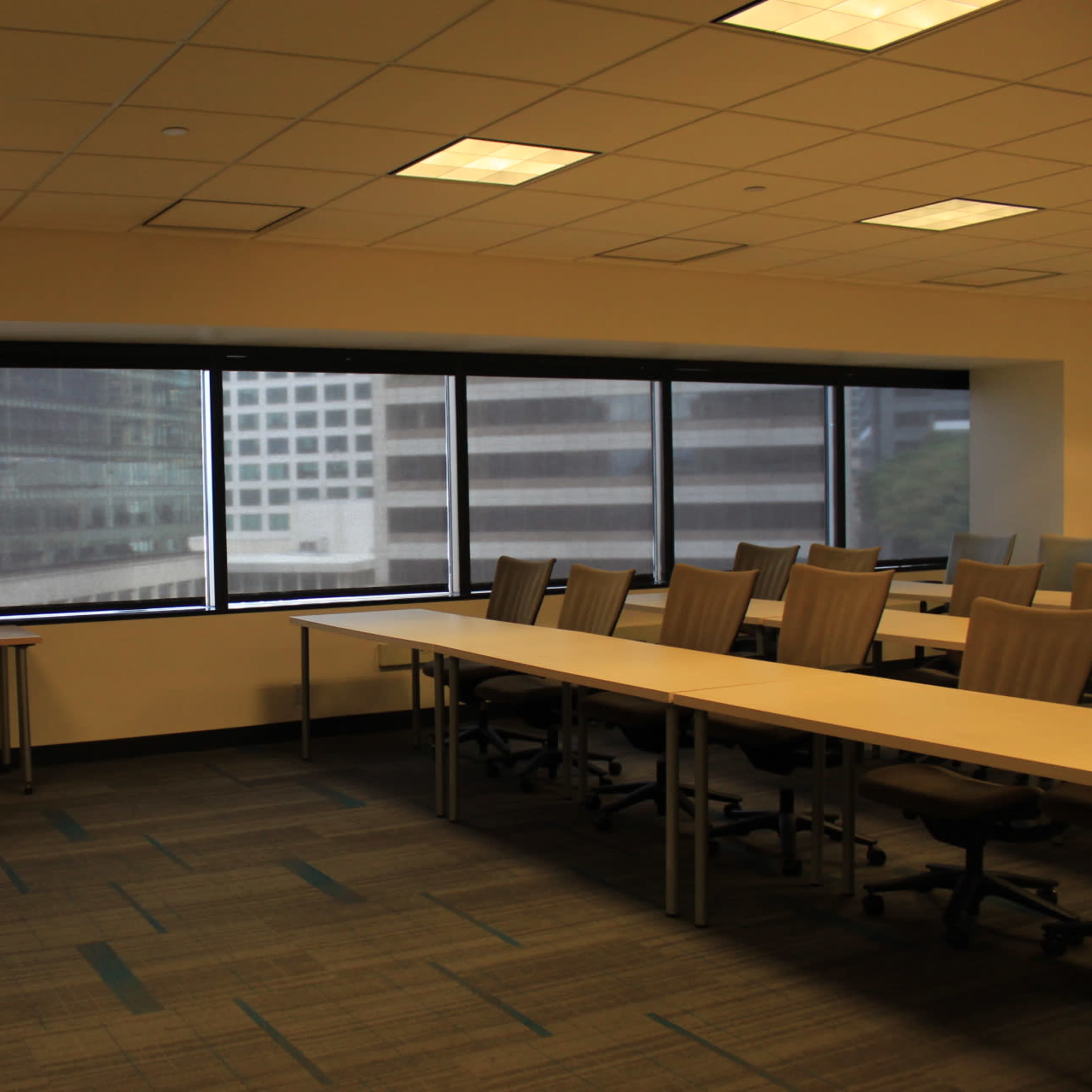 A conference room with long tables and chairs is positioned next to large windows overlooking a cityscape.