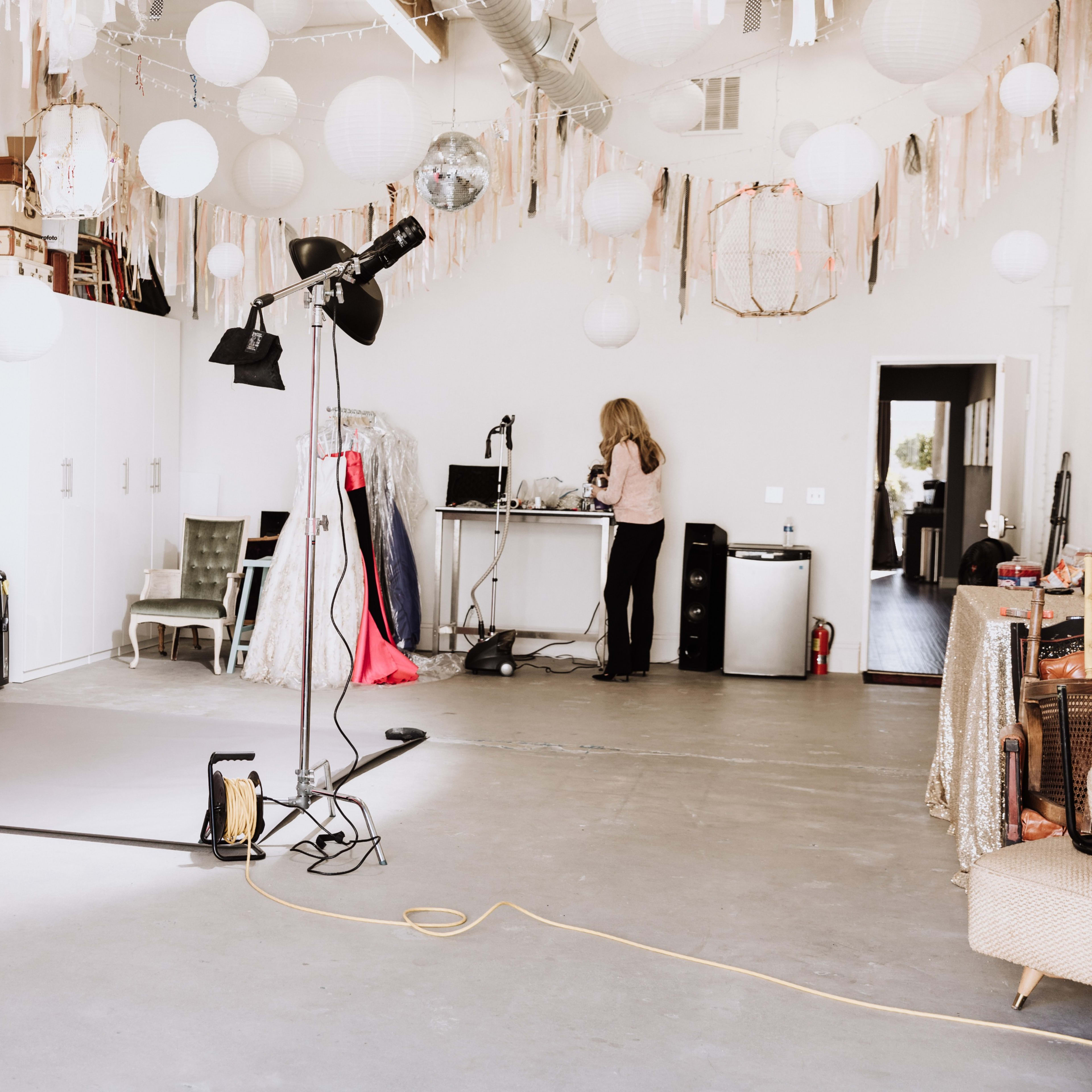 A photography studio with a gray backdrop, various decorative lanterns hanging from the ceiling, and a woman working at a desk in the background.