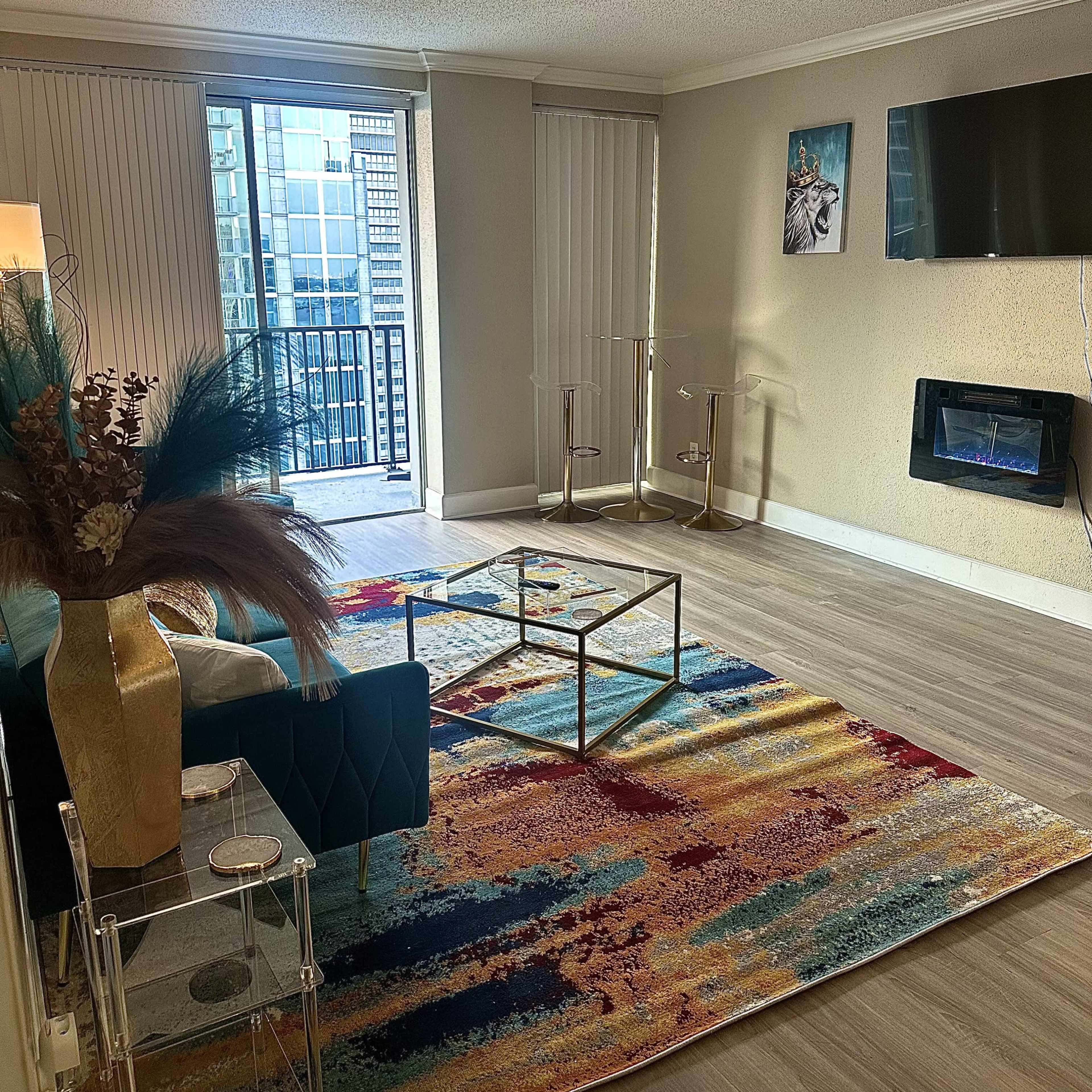 A bright living room featuring a colorful area rug, a glass coffee table, a blue armchair, and a view of a balcony through sliding glass doors.