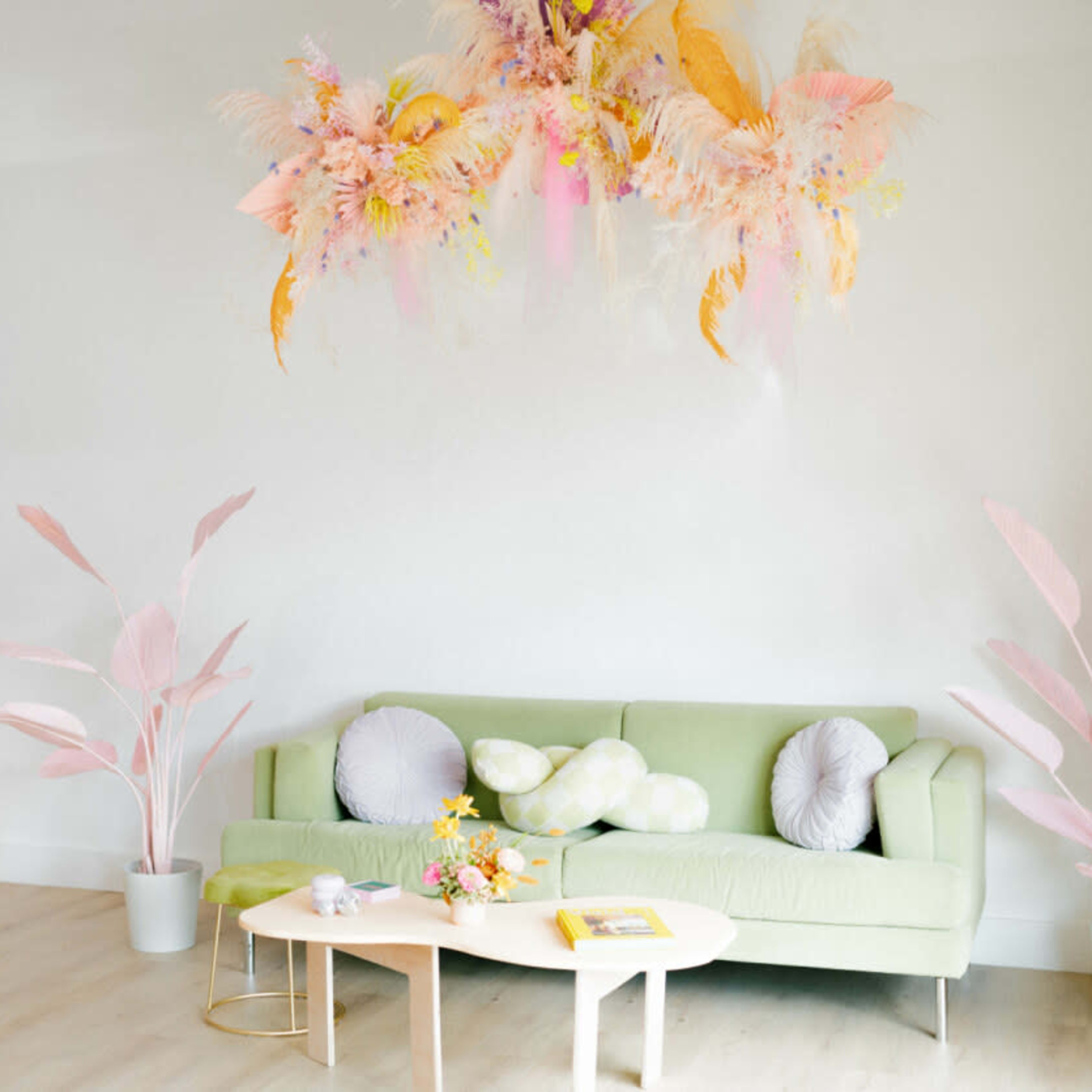 A light-filled room features a green sofa with decorative pillows, a coffee table with flowers, and pastel-colored plants and decor accents.