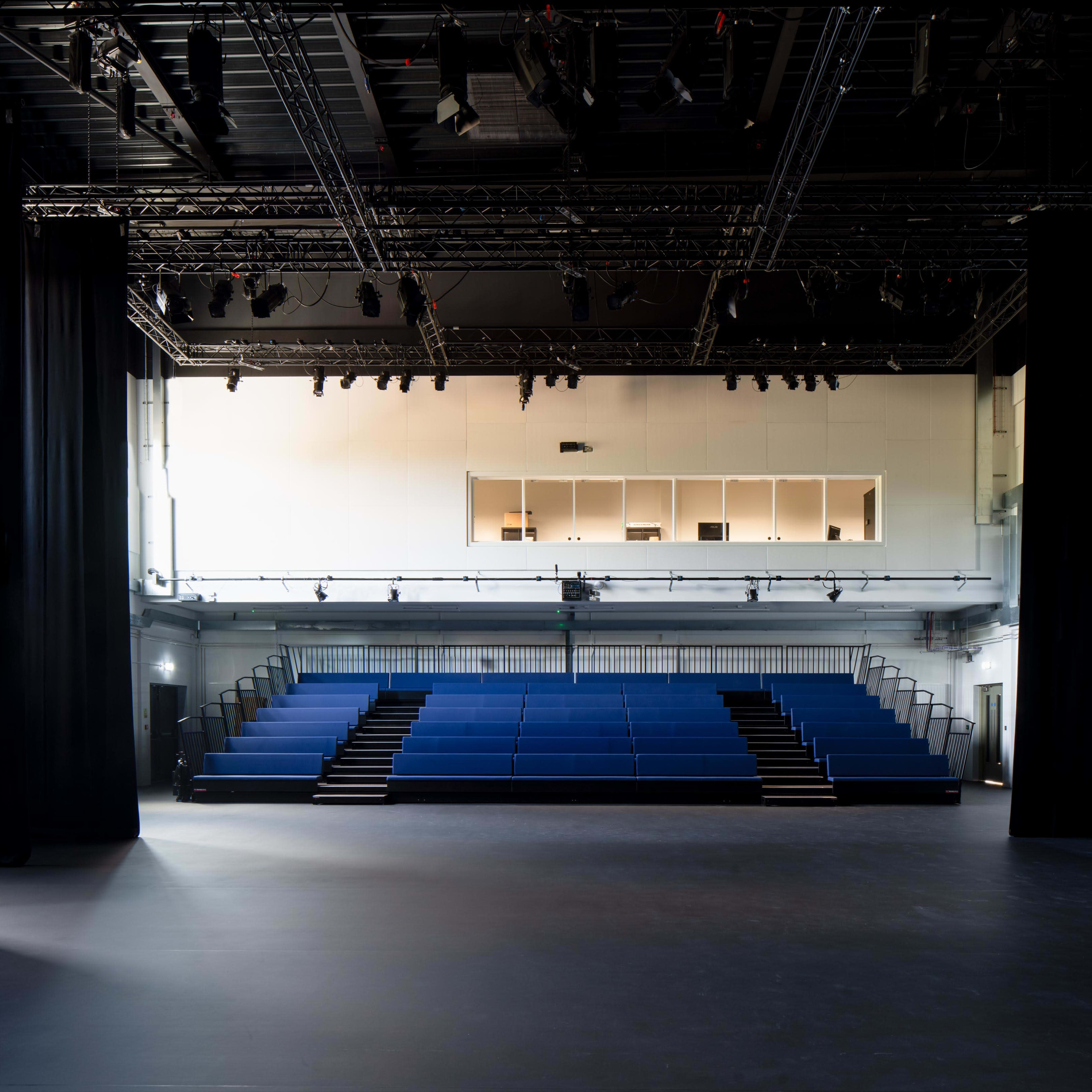 A spacious theater with blue seating arranged in a tiered formation and a well-lit stage area.