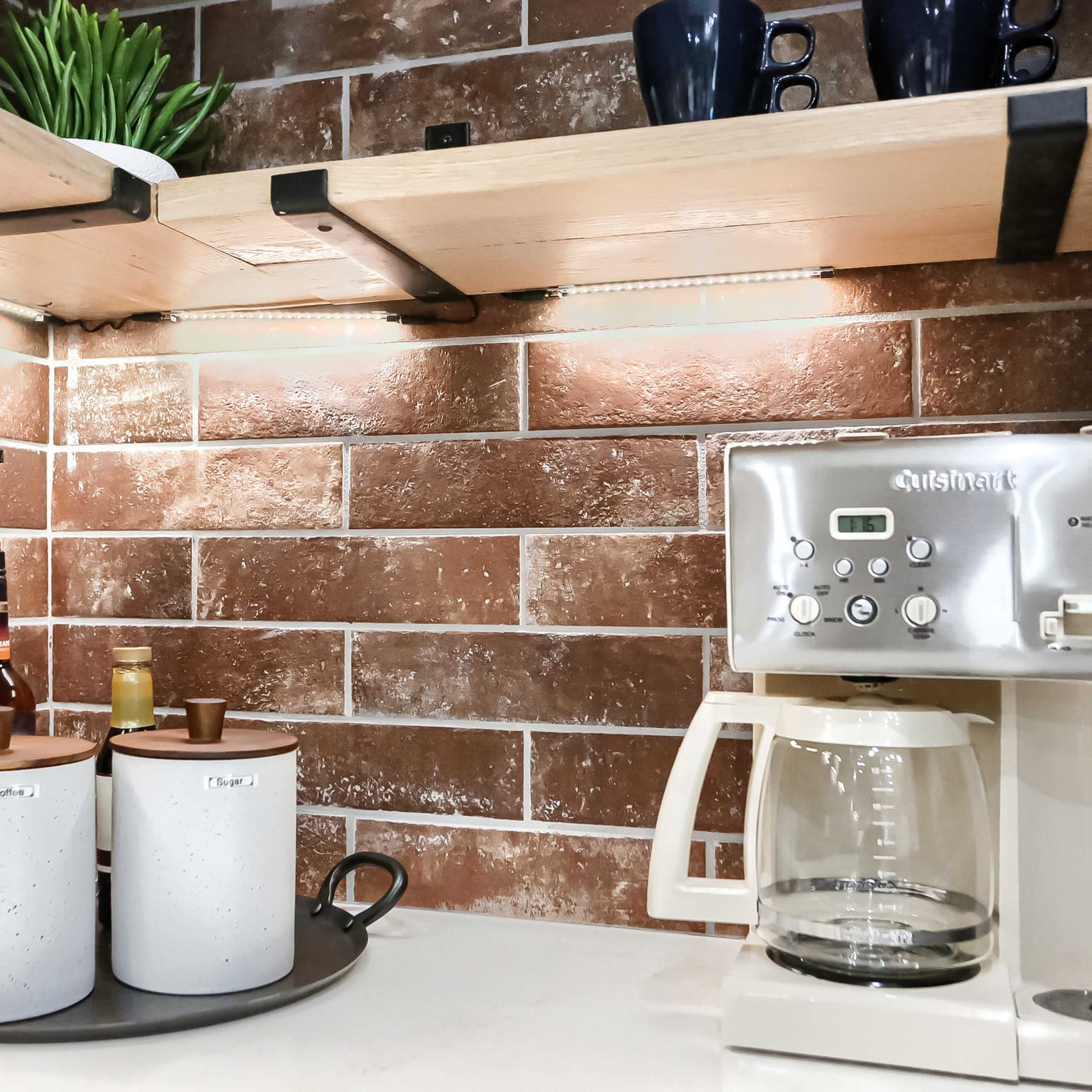A kitchen corner with a coffee maker next to round containers on a tray, all set against a brown brick wall and illuminated by shelf lights.