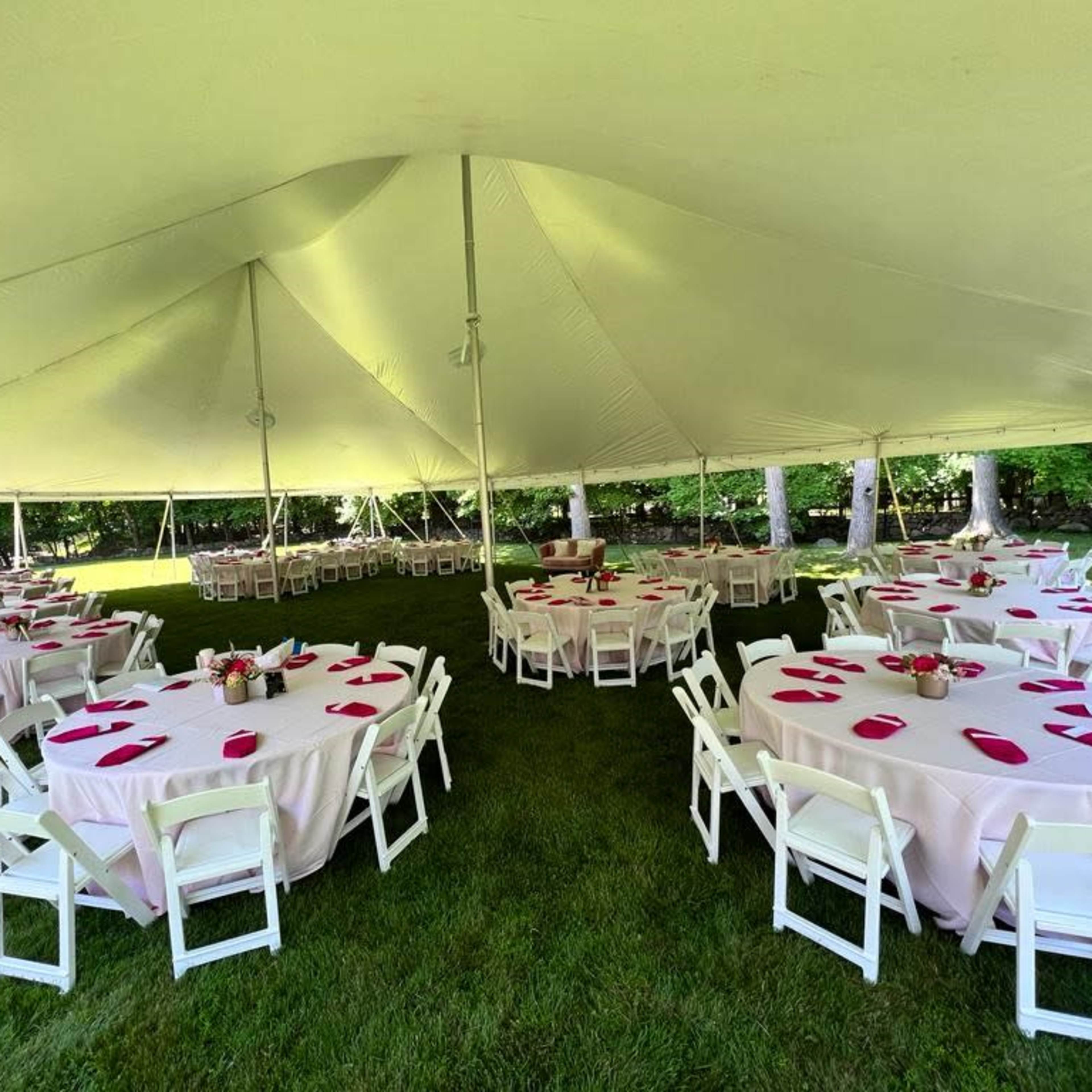 A tented outdoor event space features round tables set with pink tablecloths and bright pink placemats on a grassy area.