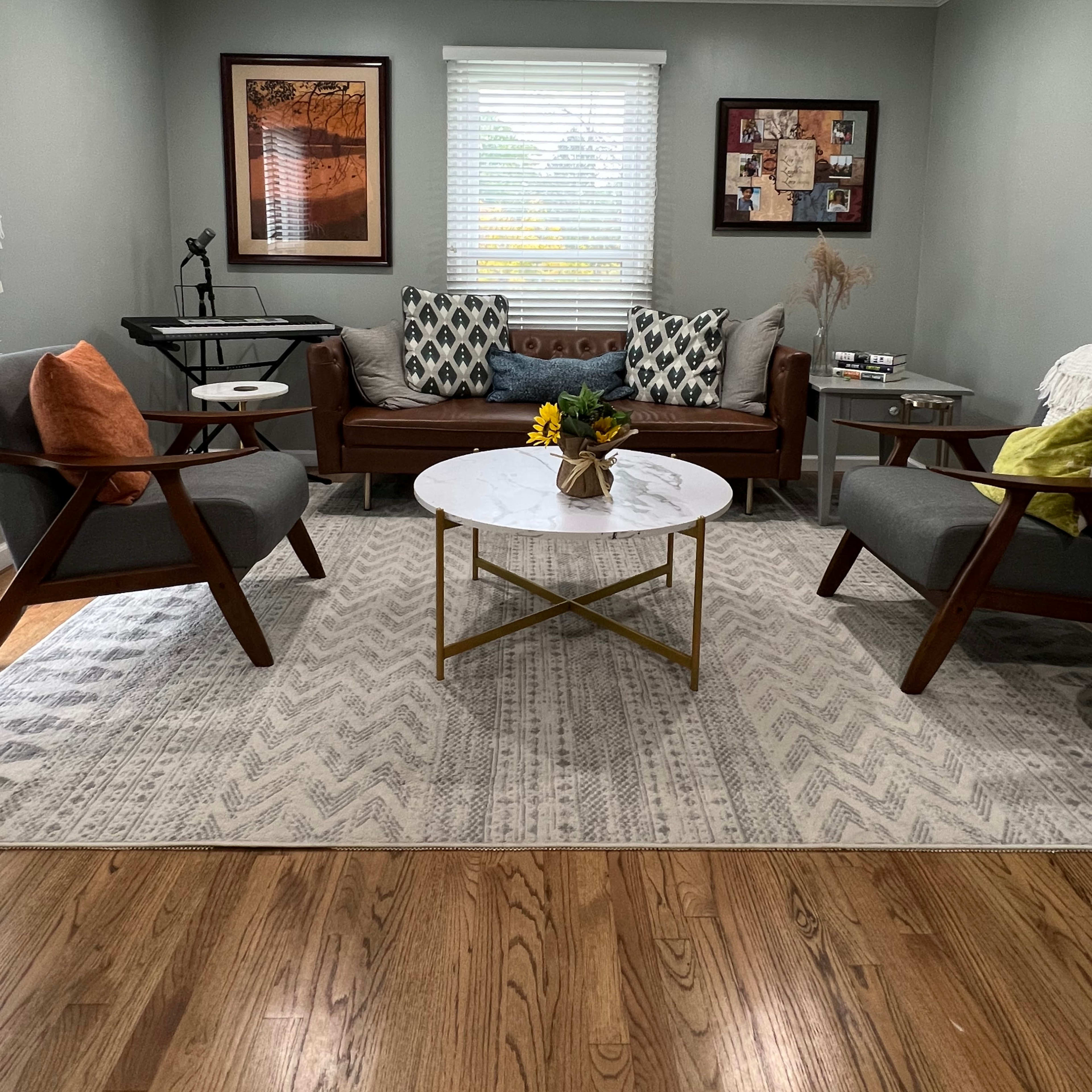 A living room with a brown leather sofa, two gray armchairs, a round coffee table with a floral arrangement, and framed artwork on the walls.