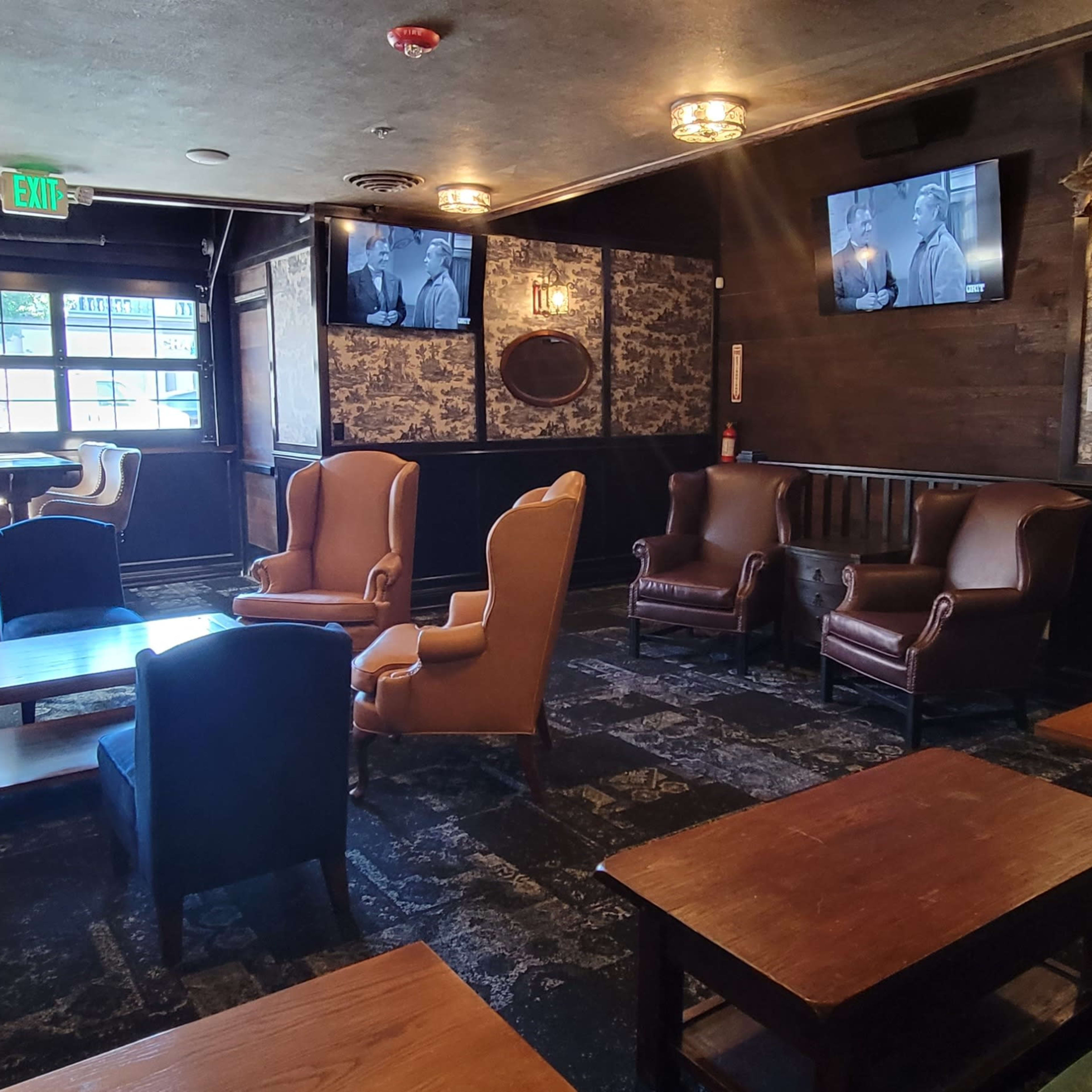 A cozy lounge area with various seating arrangements, including leather chairs and wooden tables, and large windows letting in natural light.