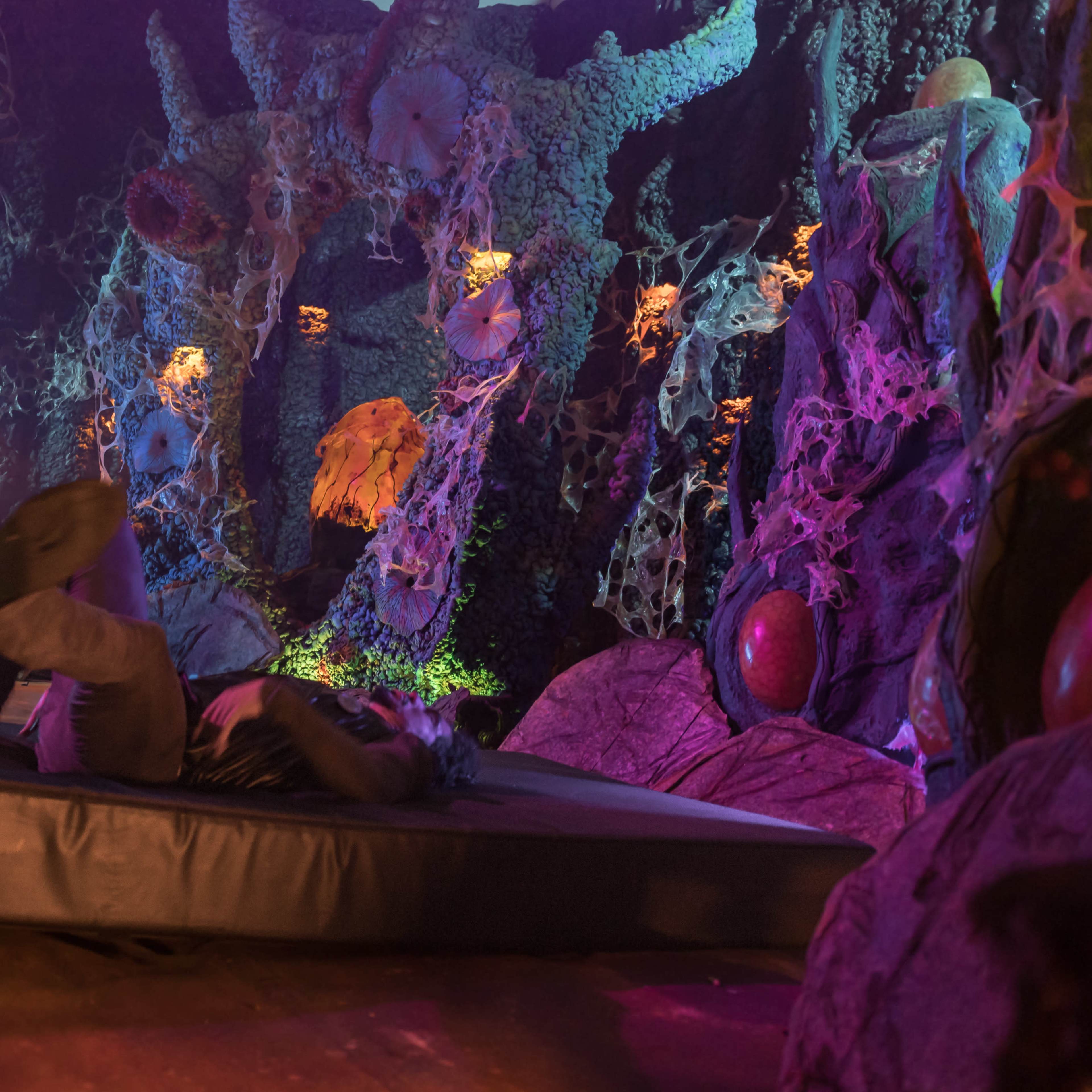 A person is lying on a padded surface surrounded by an organic-looking, illuminated environment with intricate textures and shapes.