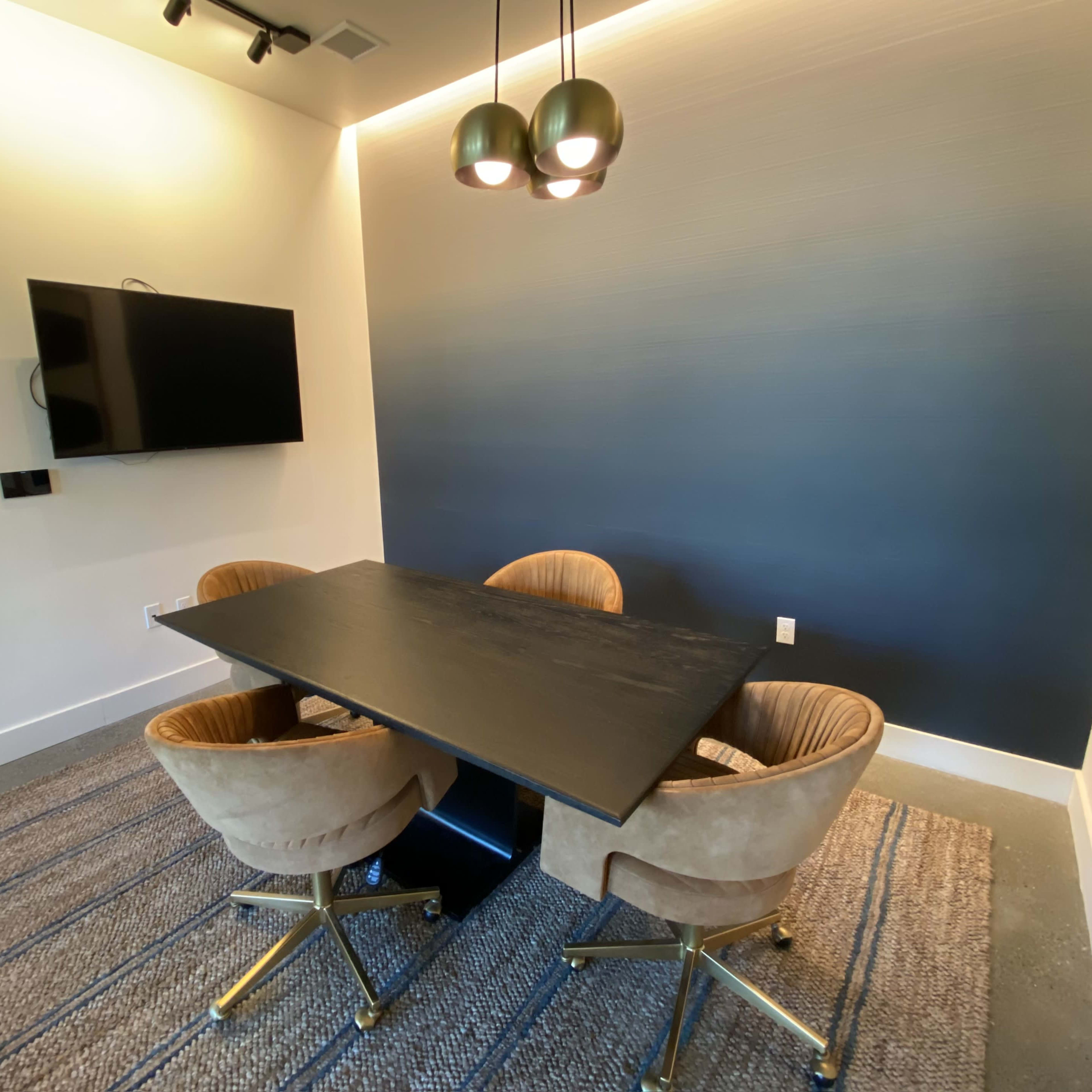 A modern meeting room features a dark table with four round chairs, a wall-mounted television, and a gradient blue wall.