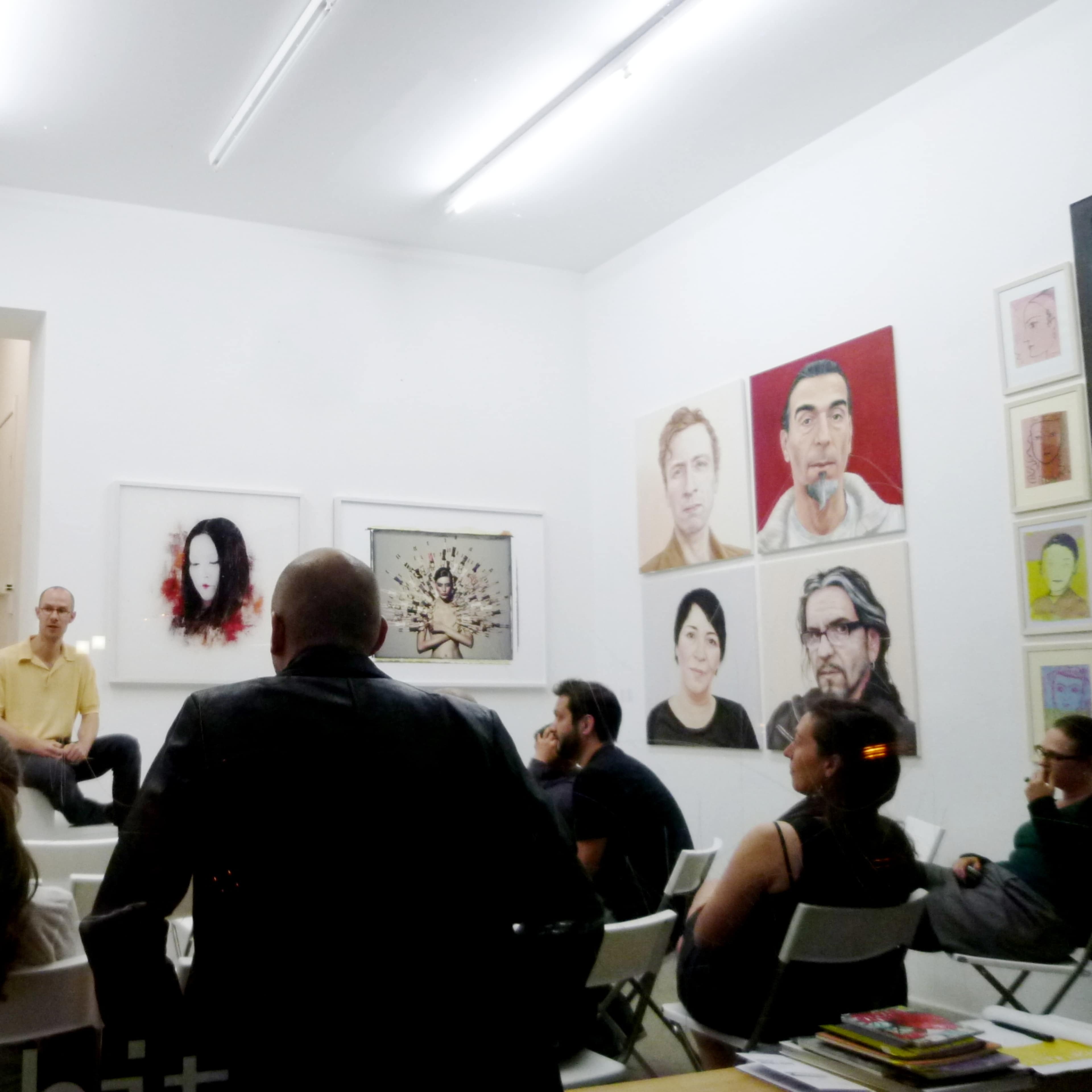 A group of people sits in white chairs facing an artist who is speaking in a gallery adorned with various artworks.