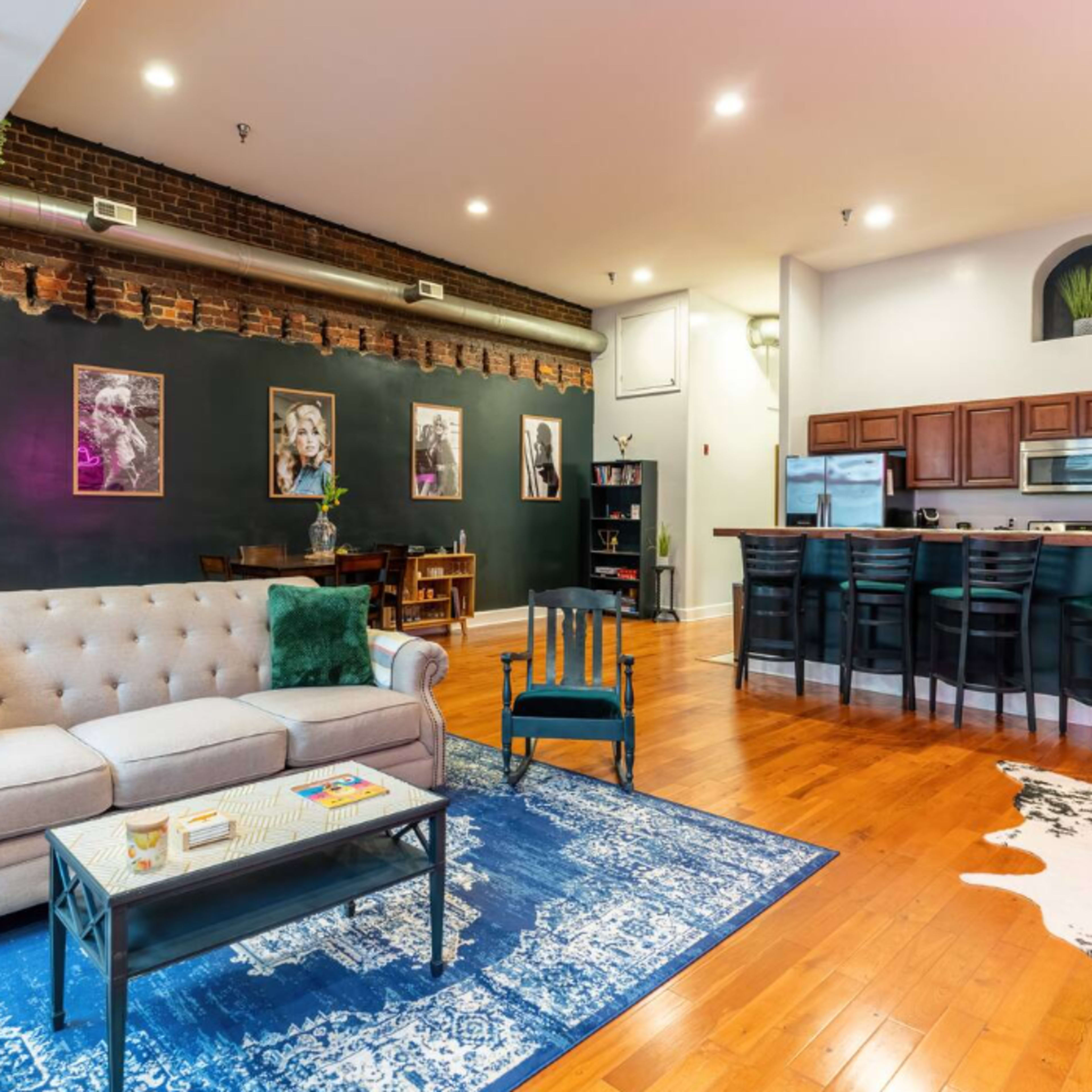 A spacious living area features a tufted sofa, an accent chair, a blue rug, and a kitchen with wooden cabinets and stainless steel appliances.