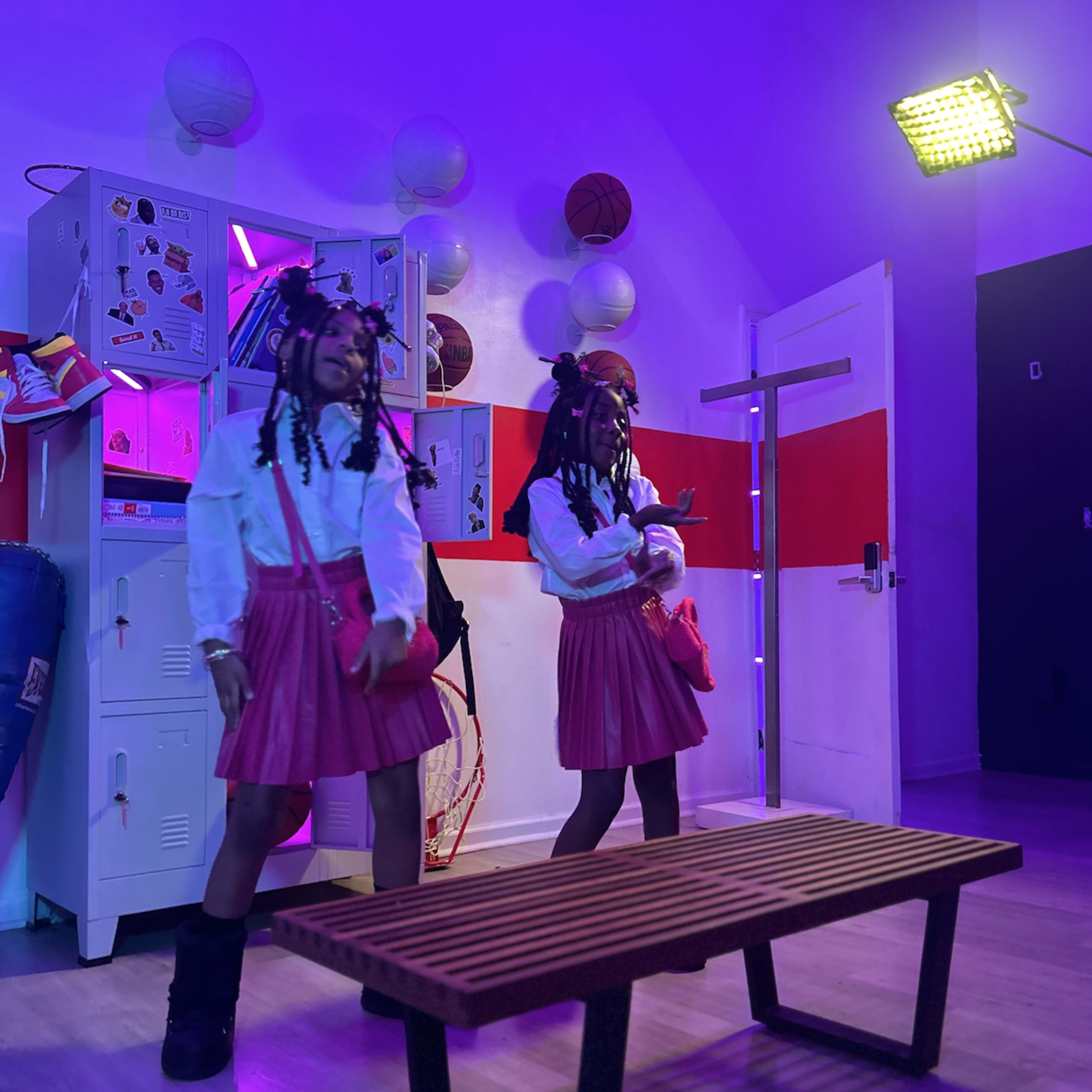 Two girls with braided hair wearing white shirts and pink skirts dance in a room illuminated by purple light, with sports equipment and lockers in the background.
