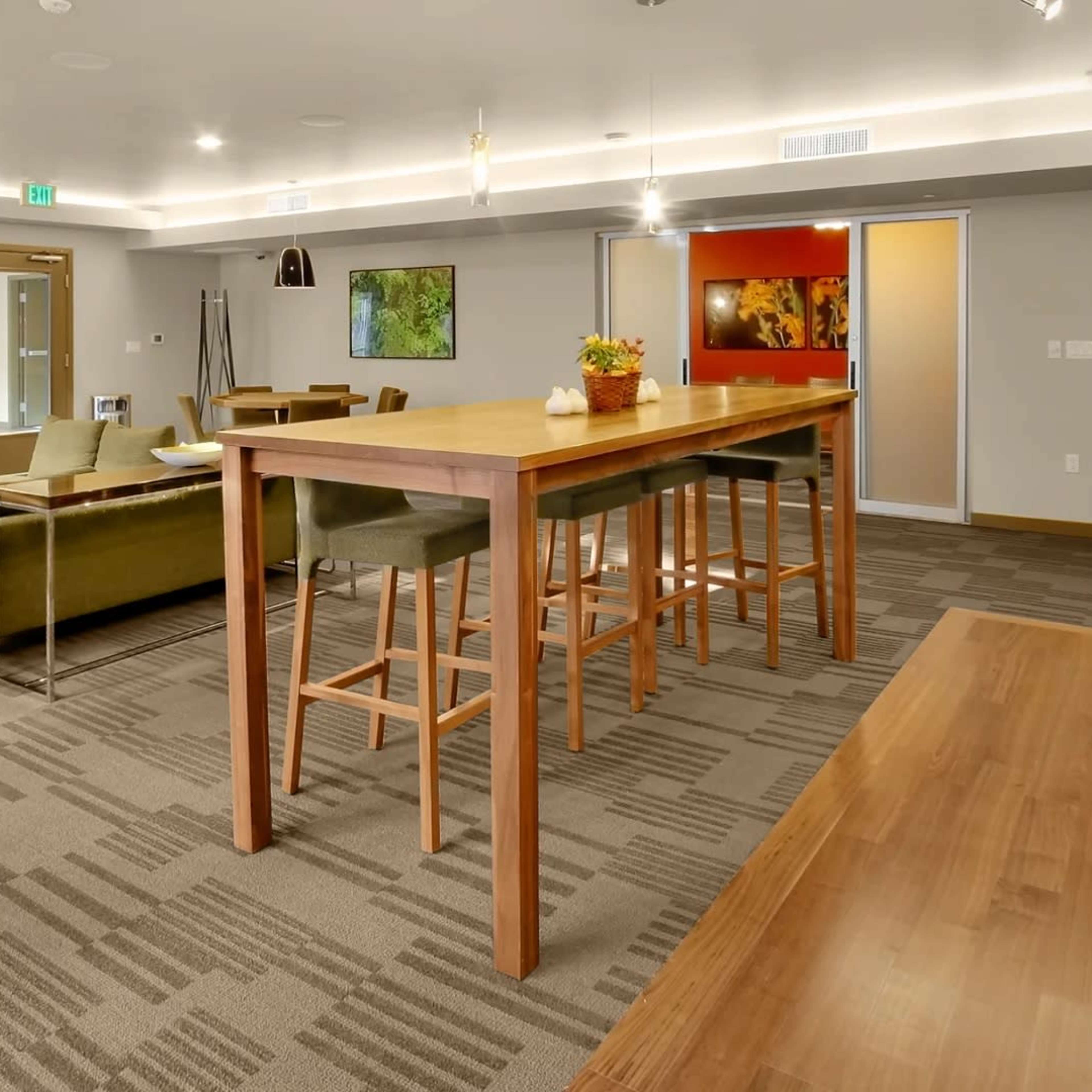 A modern common area featuring a large wooden table surrounded by high stools, a green couch, and a television on the wall.