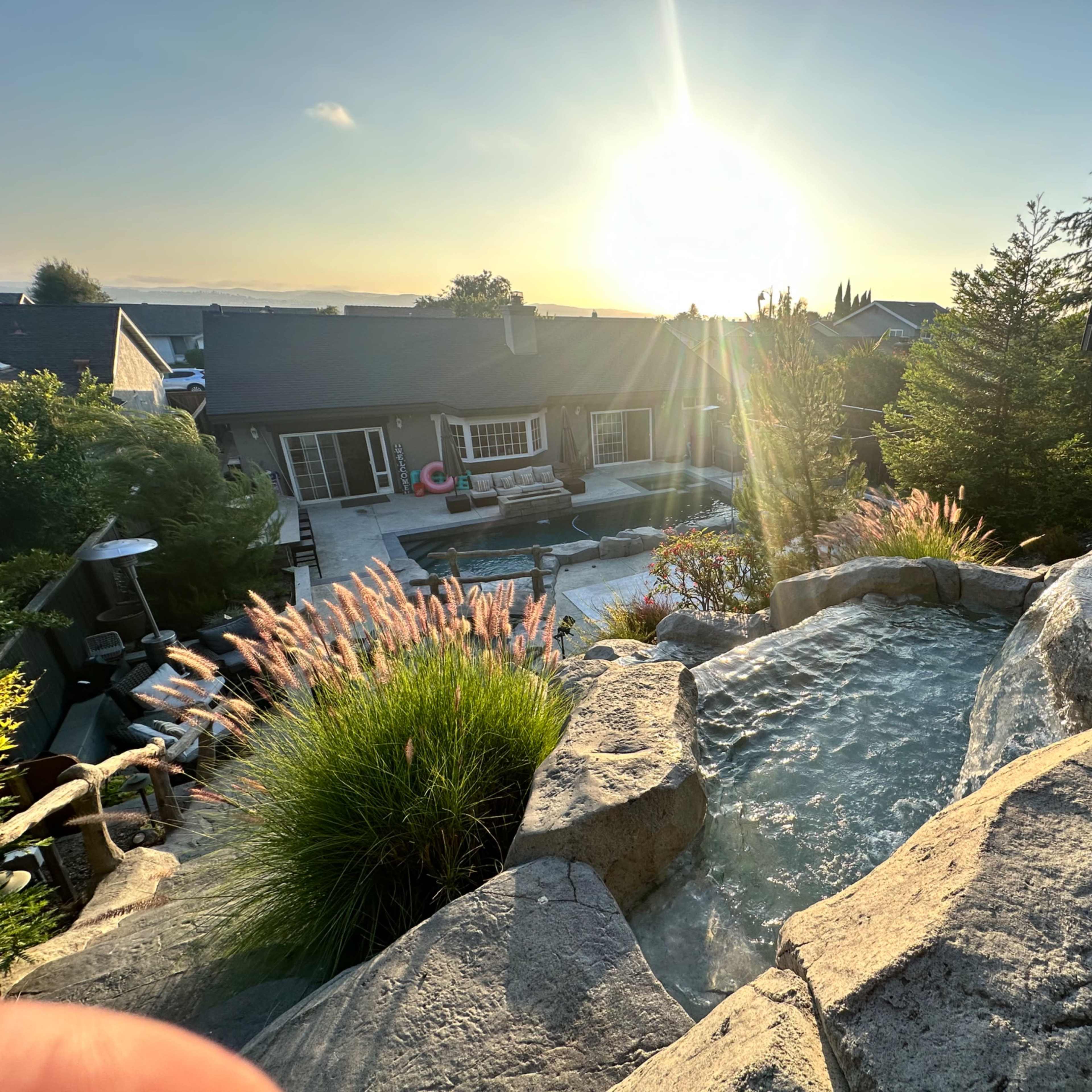 A stone hot tub and landscaping with a view of a house and a swimming pool illuminated by the sunset.