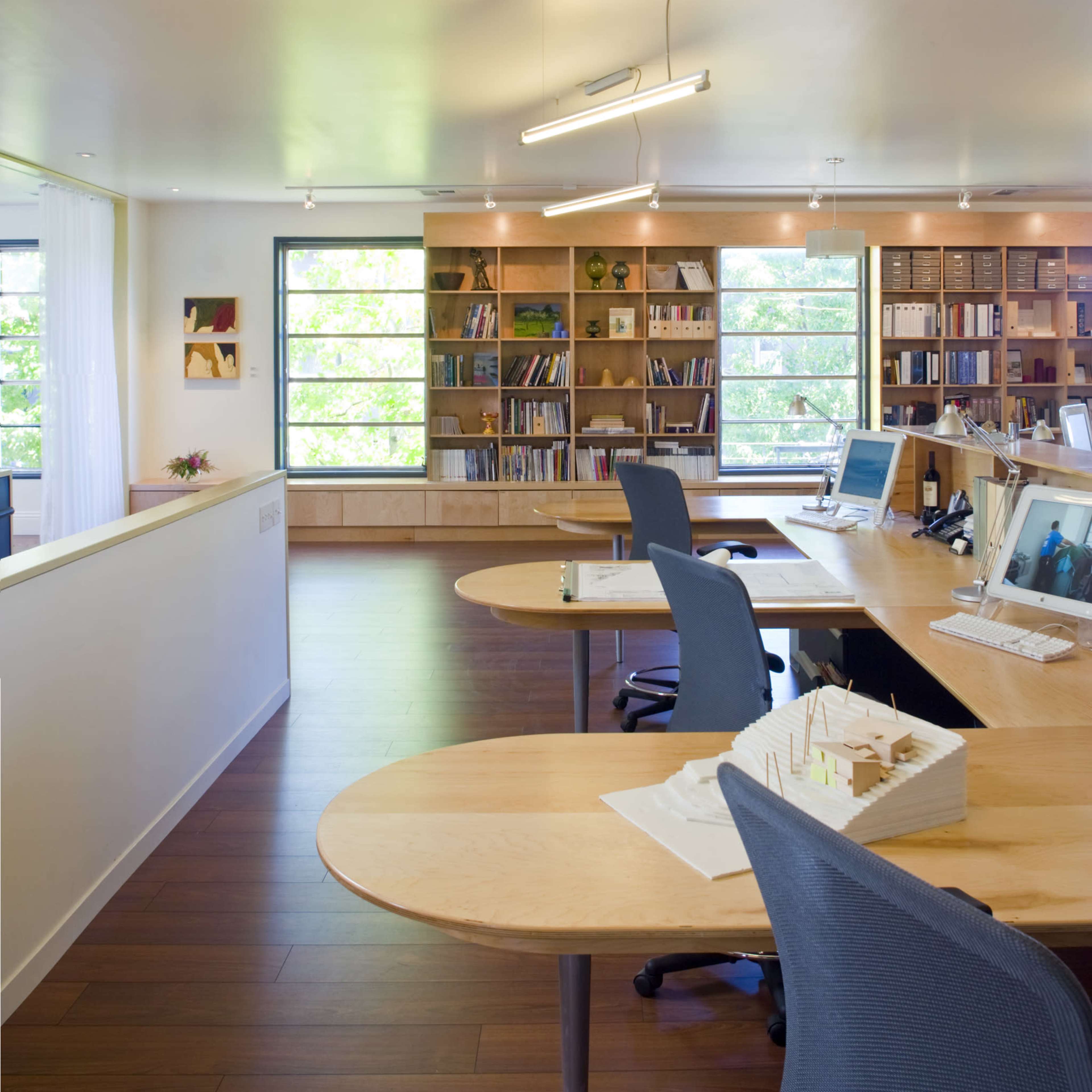 A modern office space with wooden desks, ergonomic chairs, and shelves filled with books and decorative items.