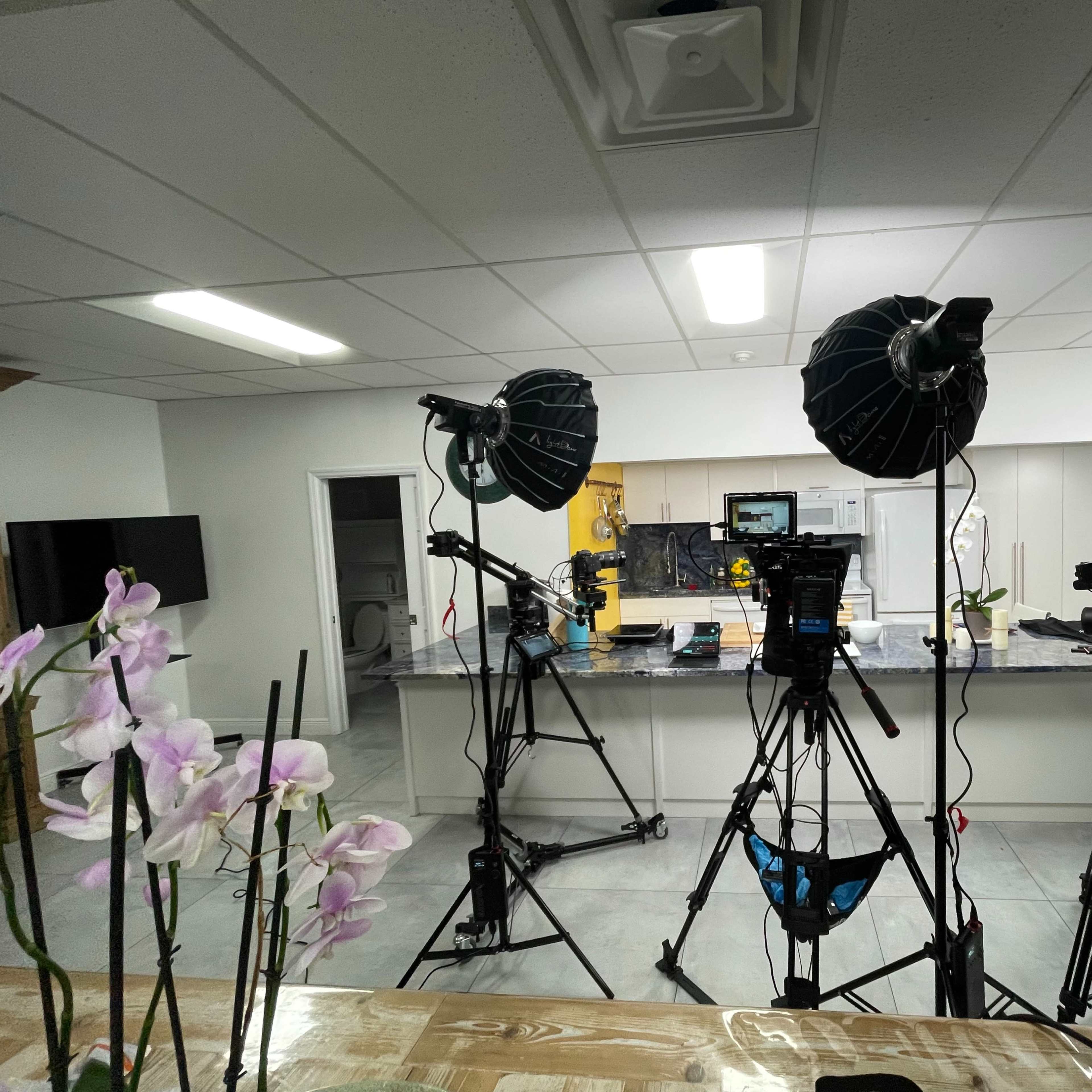 A well-lit kitchen set up for filming, featuring multiple cameras and lighting equipment in front of a counter.