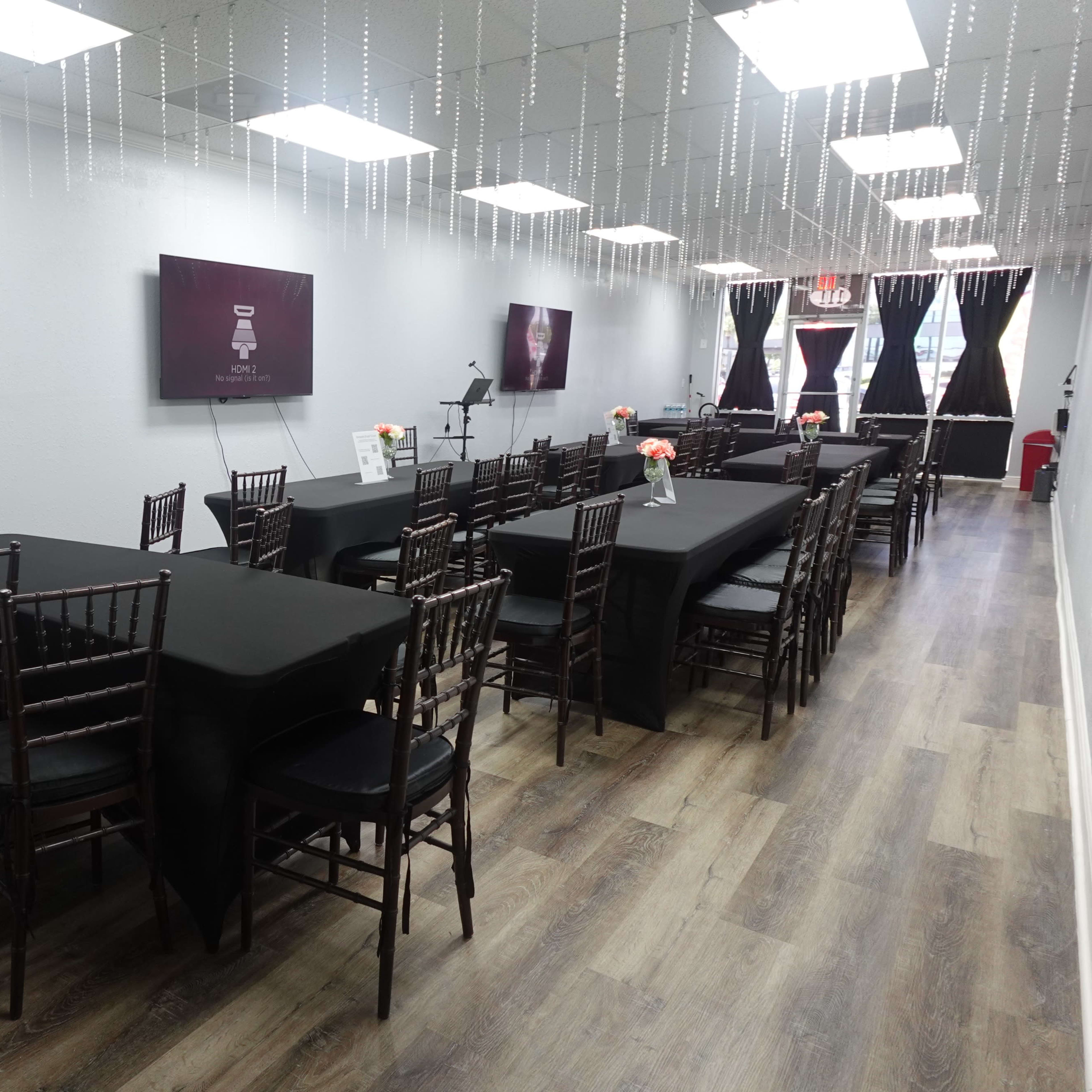A long event space with black tablecloths, chiavari chairs, and decorative lighting hanging from the ceiling.