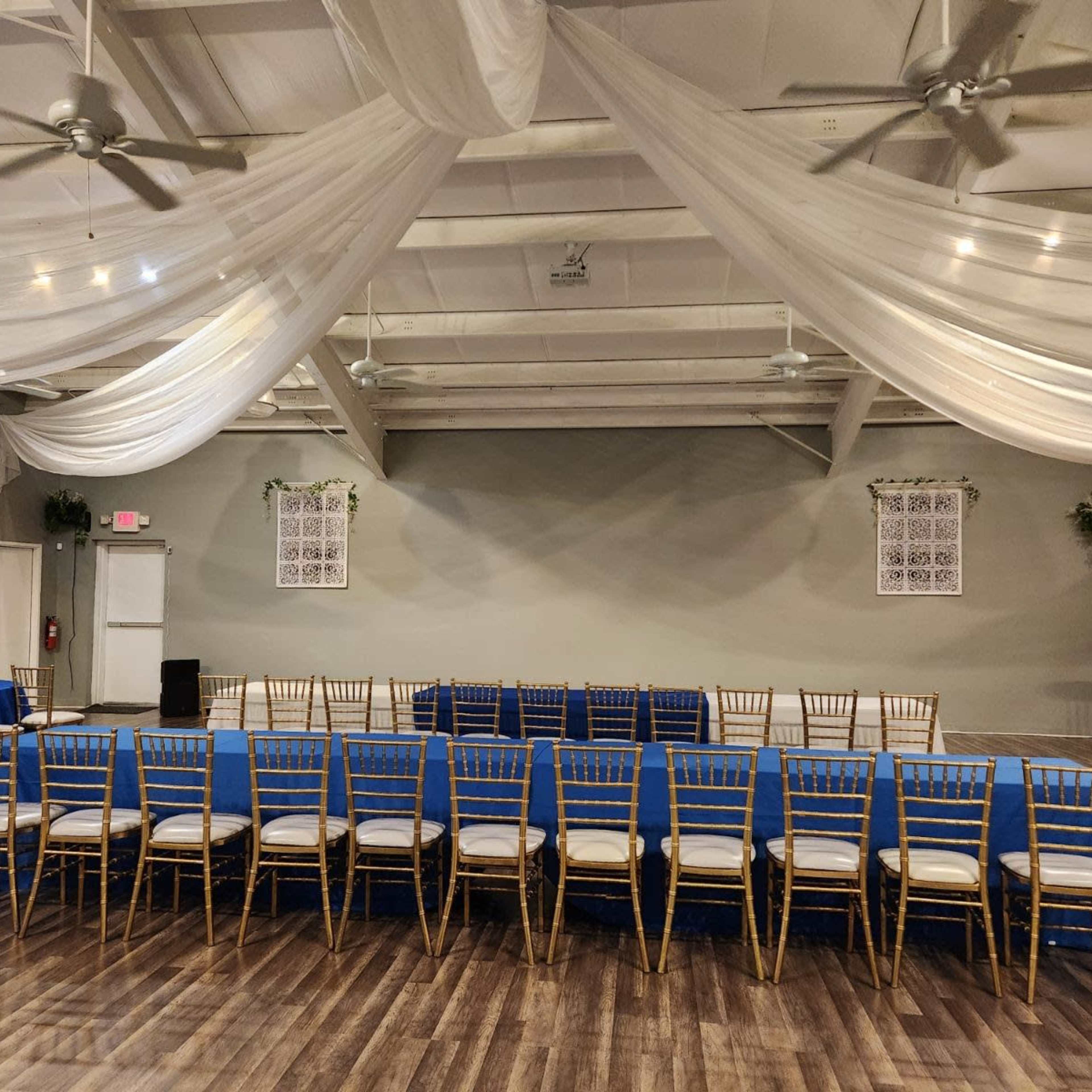 A spacious indoor venue with blue-covered tables arranged in a layout and draped fabric overhead.