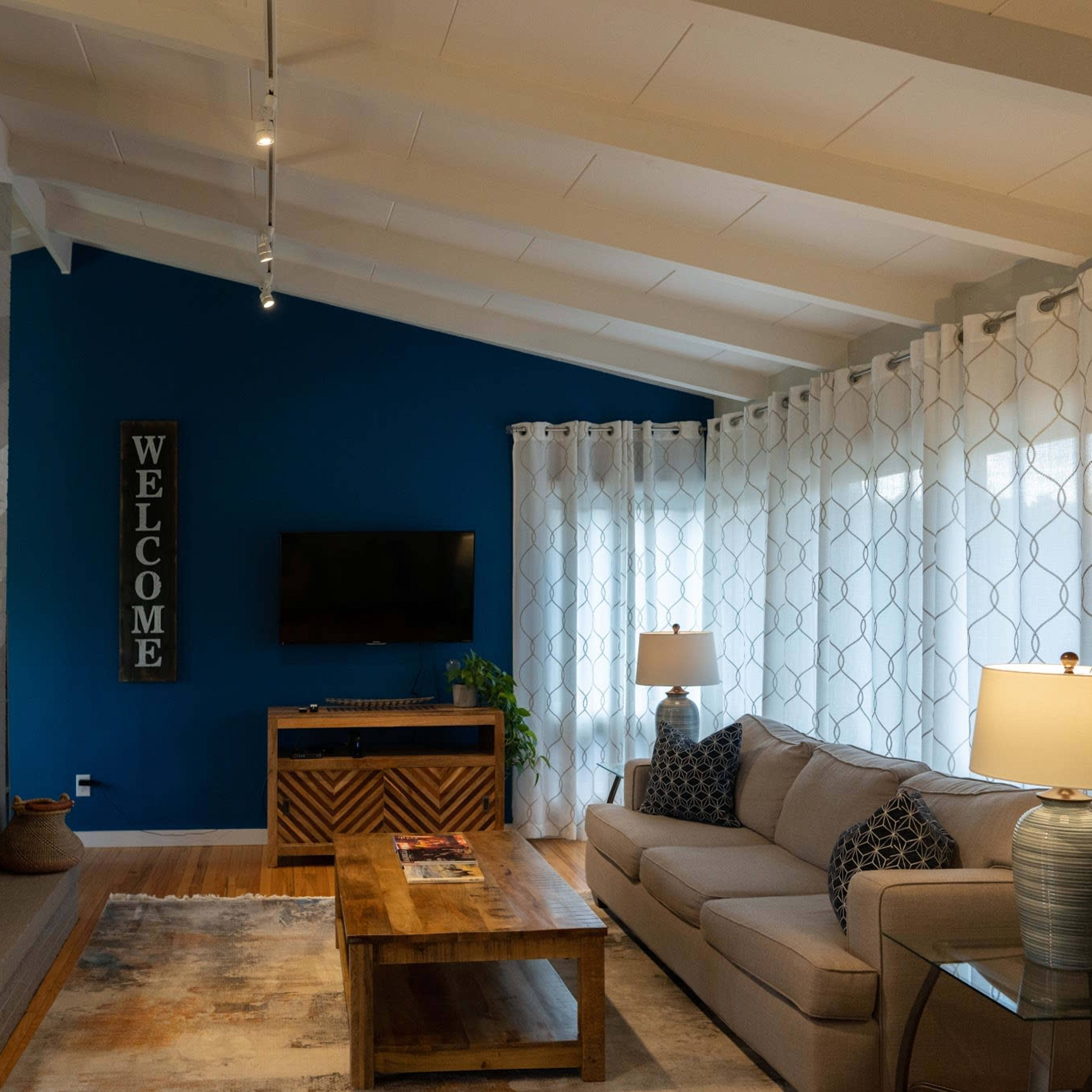 A modern living room features a gray sofa, wooden coffee table, and a blue accent wall with a mounted TV.