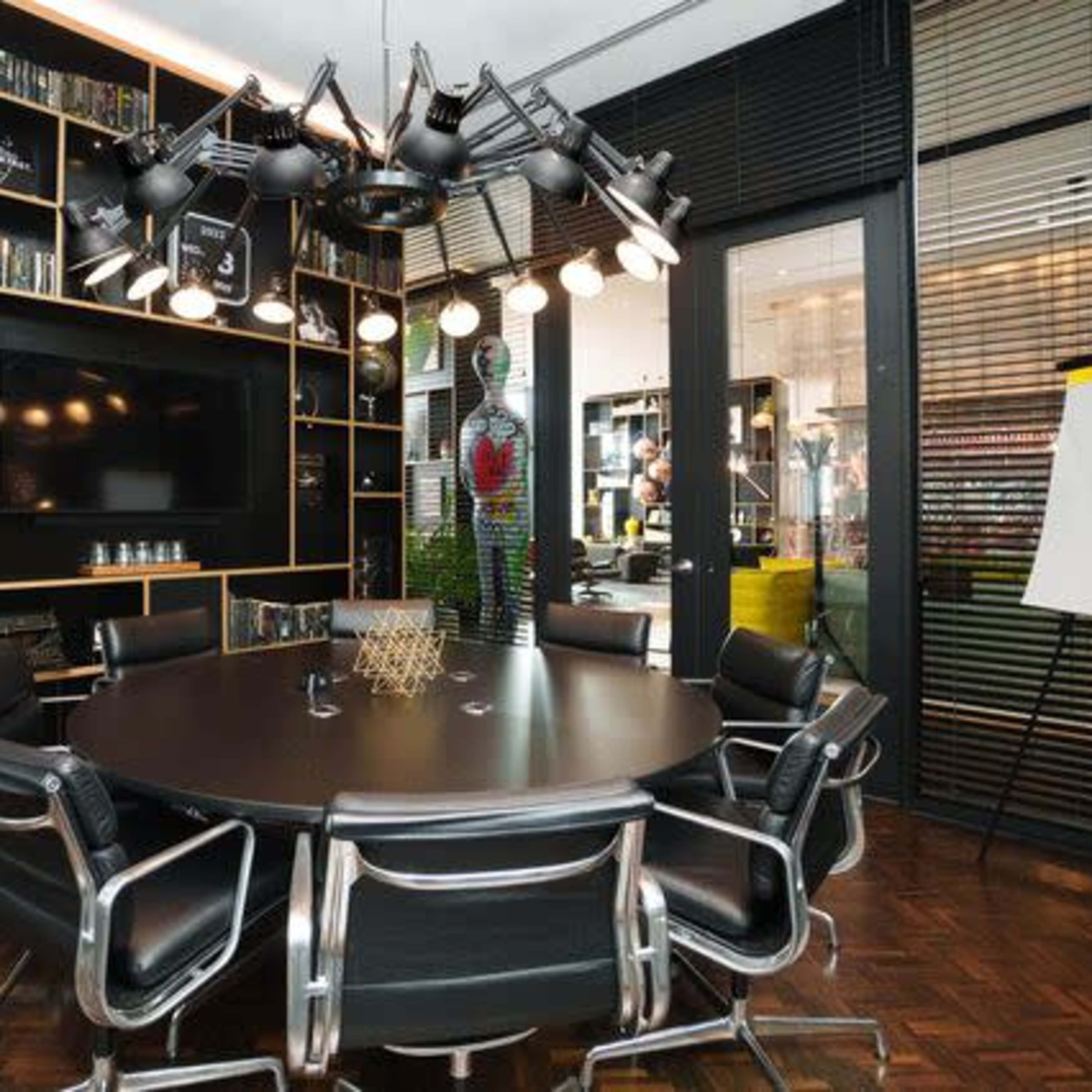 A modern conference room features a round table surrounded by sleek black chairs, a large TV, and wall-mounted bookshelves filled with various items.