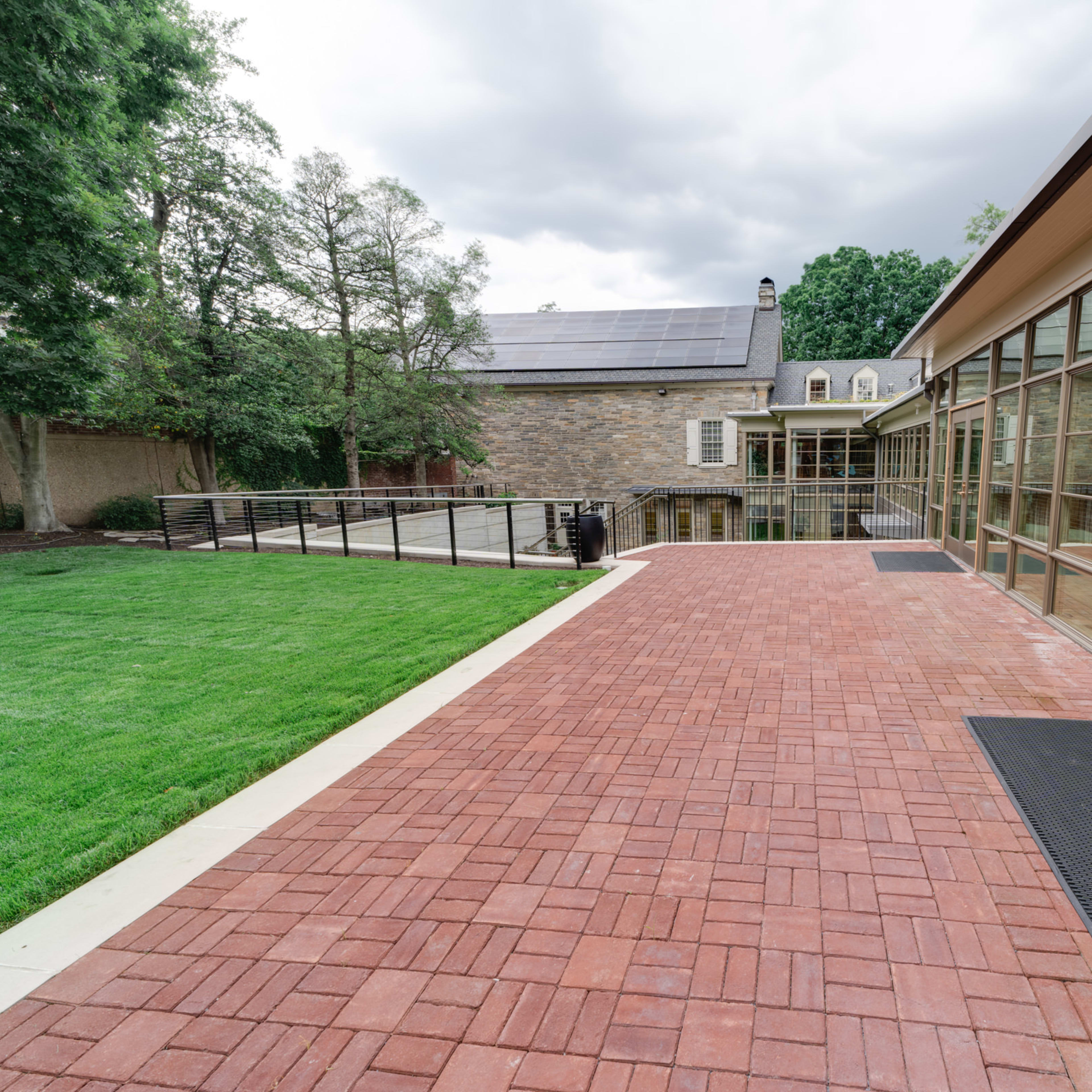 A brick patio area adjacent to a modern glass building and a stone structure, with green grass and trees in the background under a cloudy sky.