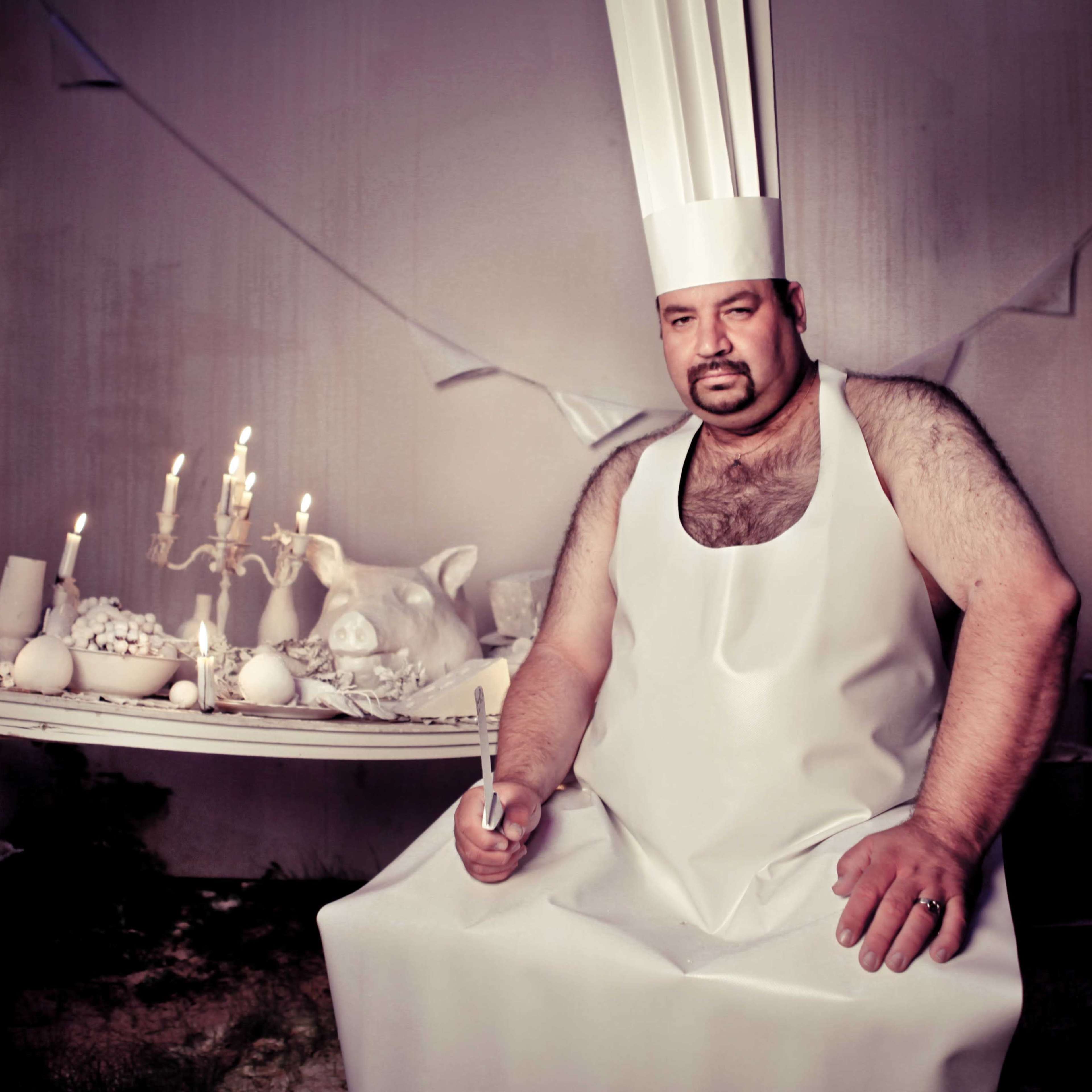 A man in a white chef's hat and apron sits at a table adorned with candles and a pig's head, surrounded by a minimalist, white-themed interior.
