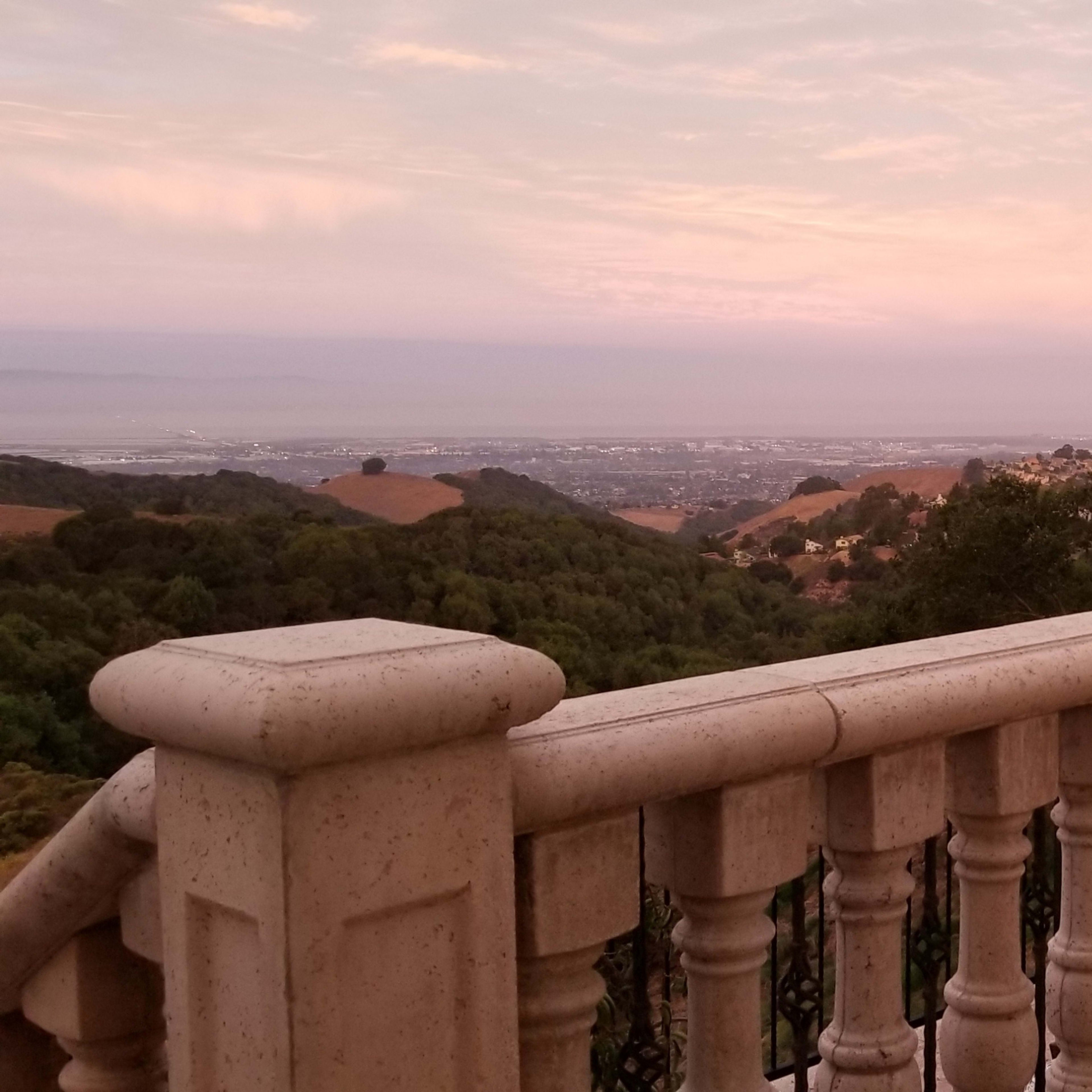 A stone balcony overlooks a vast valley at dawn, with rolling hills and a distant city visible in the background.