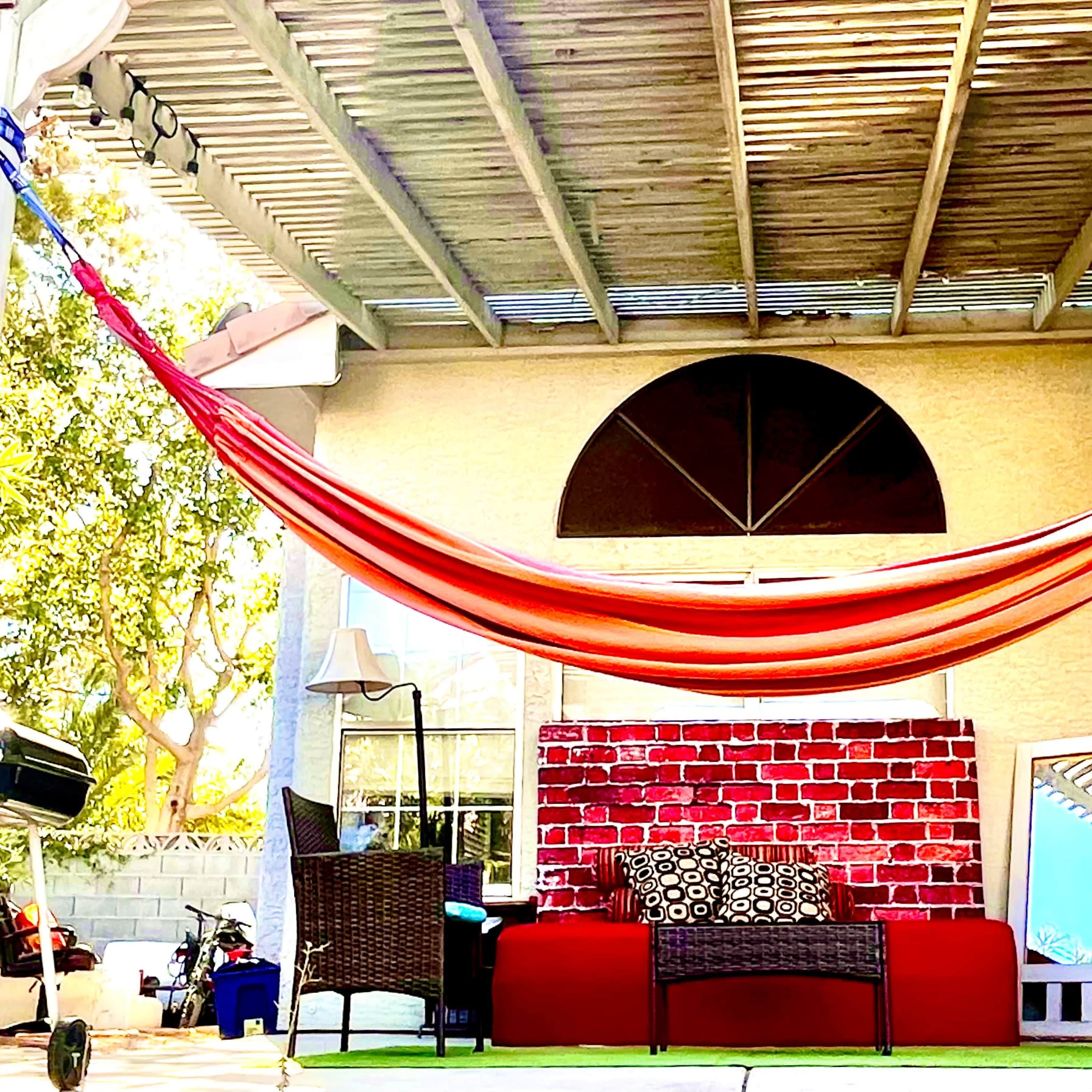 A patio area featuring a red and orange hammock suspended over a red couch, with wicker chairs and a mirror against a textured wall.