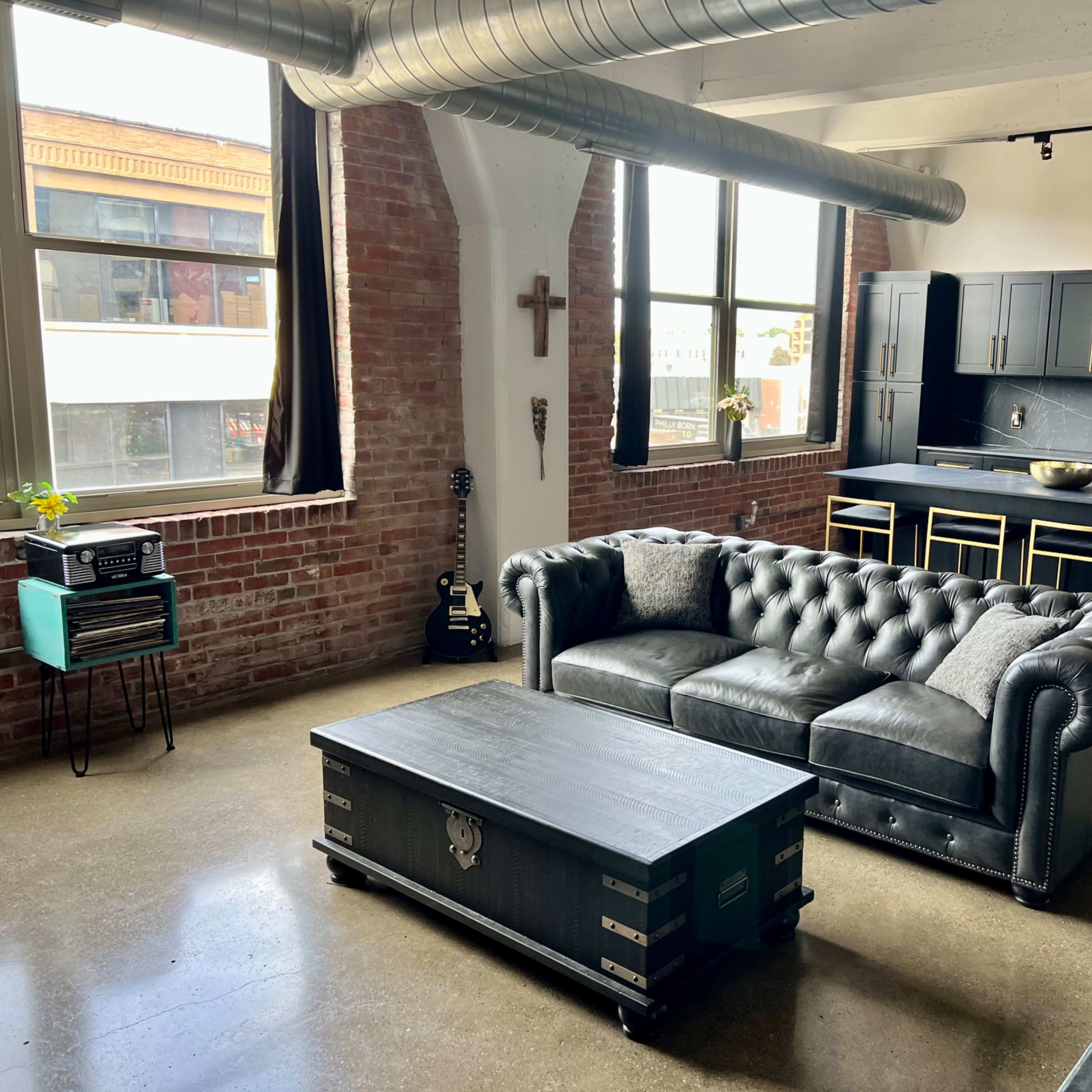A modern industrial-style living space with a black leather chesterfield sofa, a coffee table, and a kitchen area featuring dark cabinetry and metallic accents.