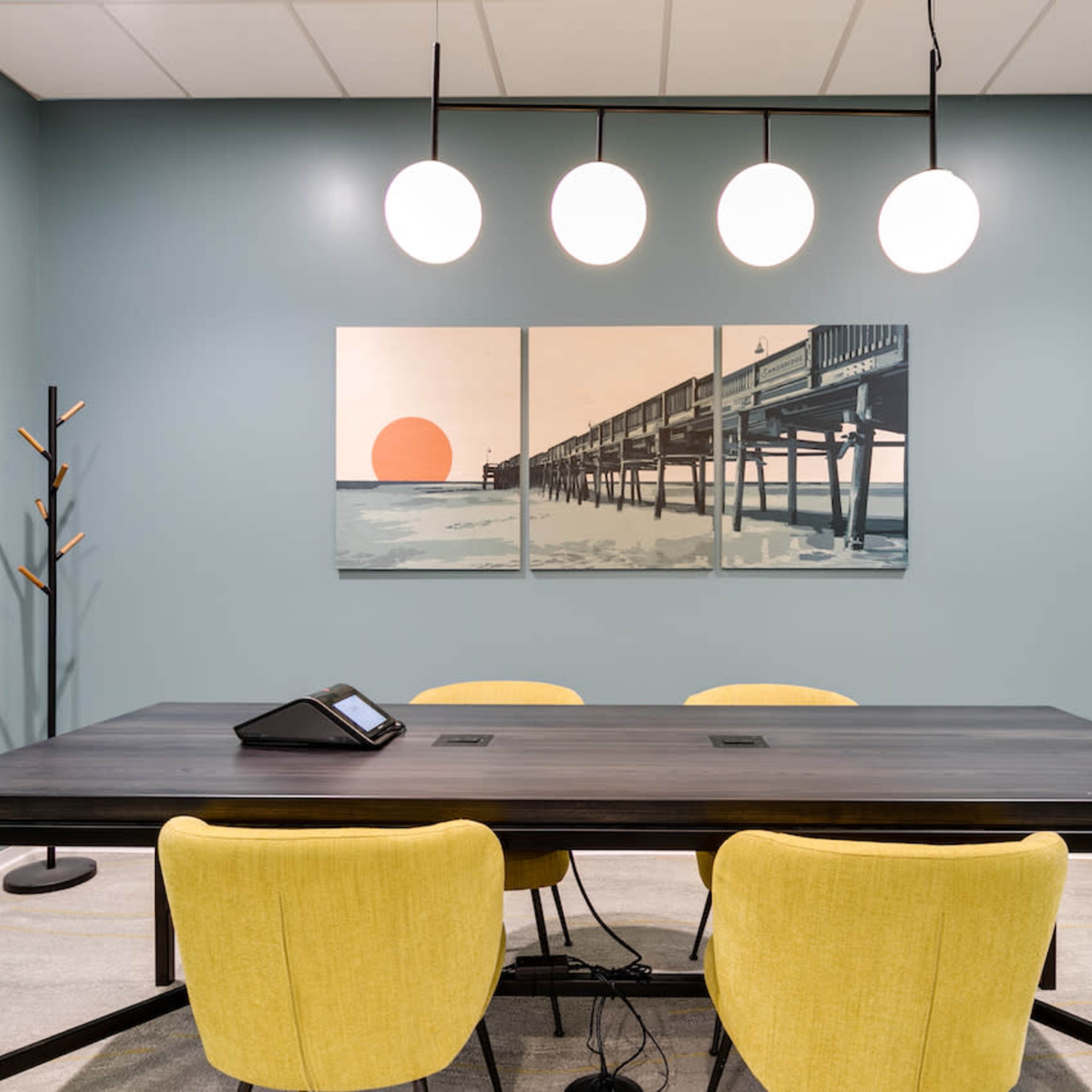 A meeting room features a large black table surrounded by yellow chairs, with modern lighting above and a mural of a pier and sunset on the wall.