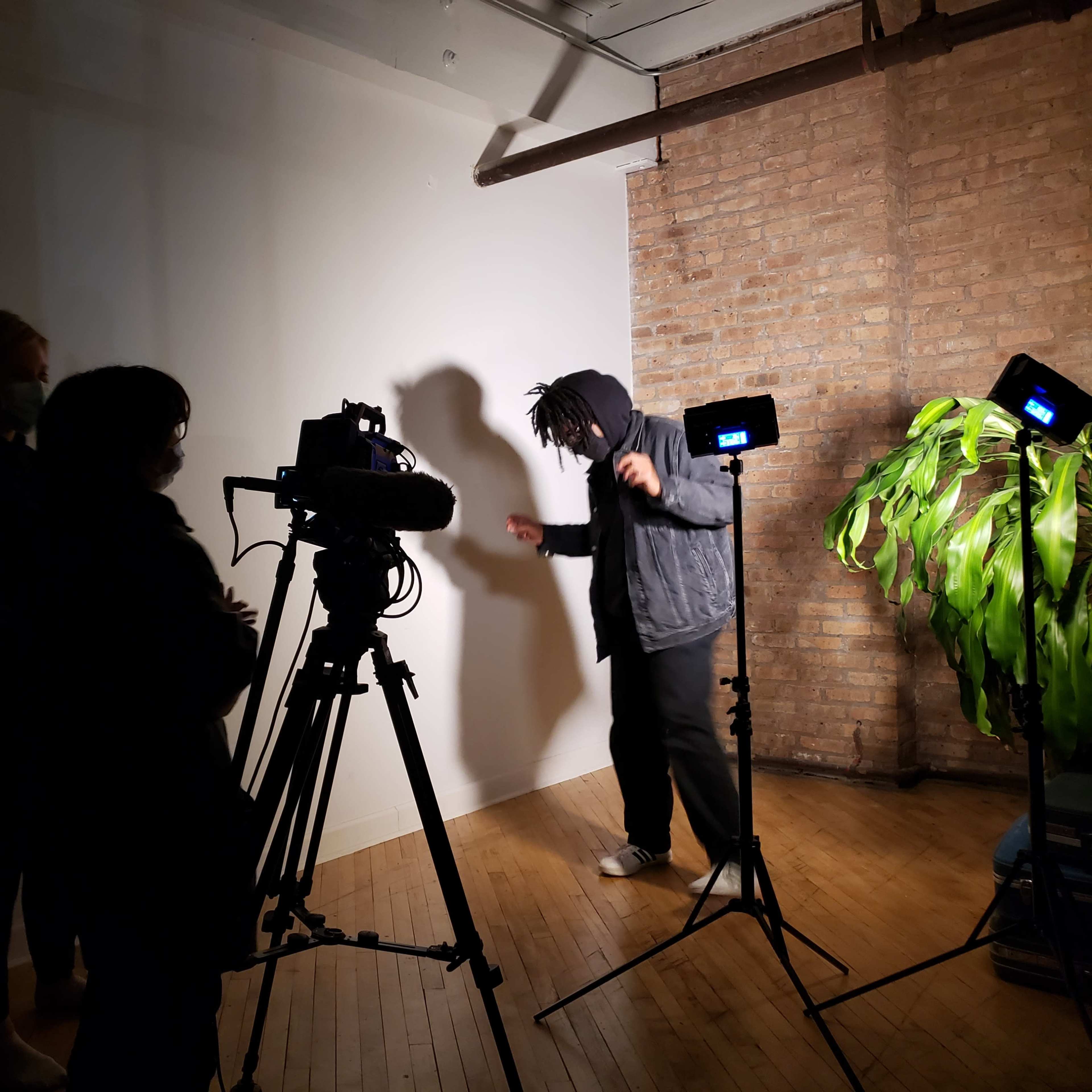 A person in a hoodie performs in front of a camera and two lights, while two others observe in a studio setting.