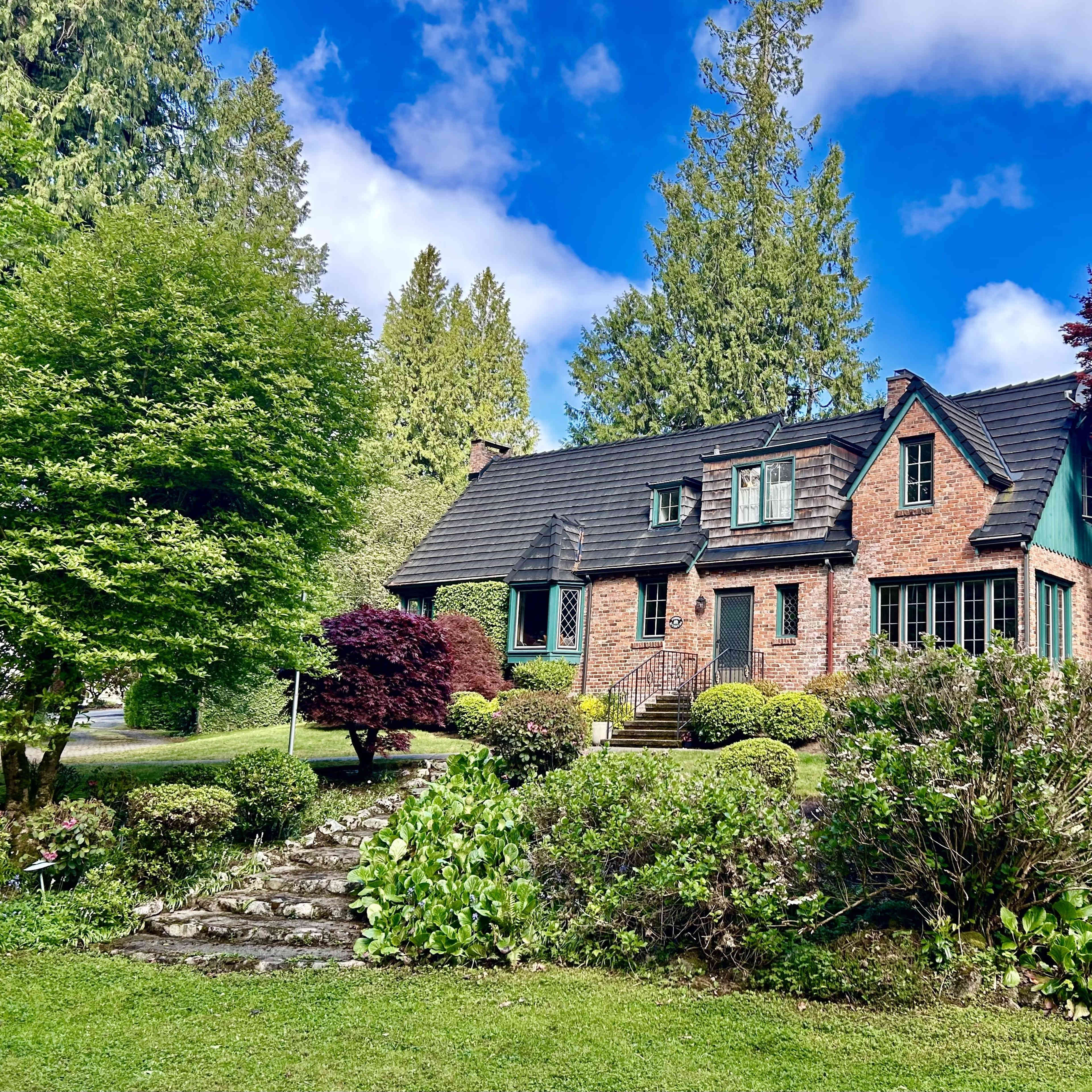 A brick house with a steep roof is surrounded by neatly trimmed bushes and trees under a blue sky with scattered clouds.