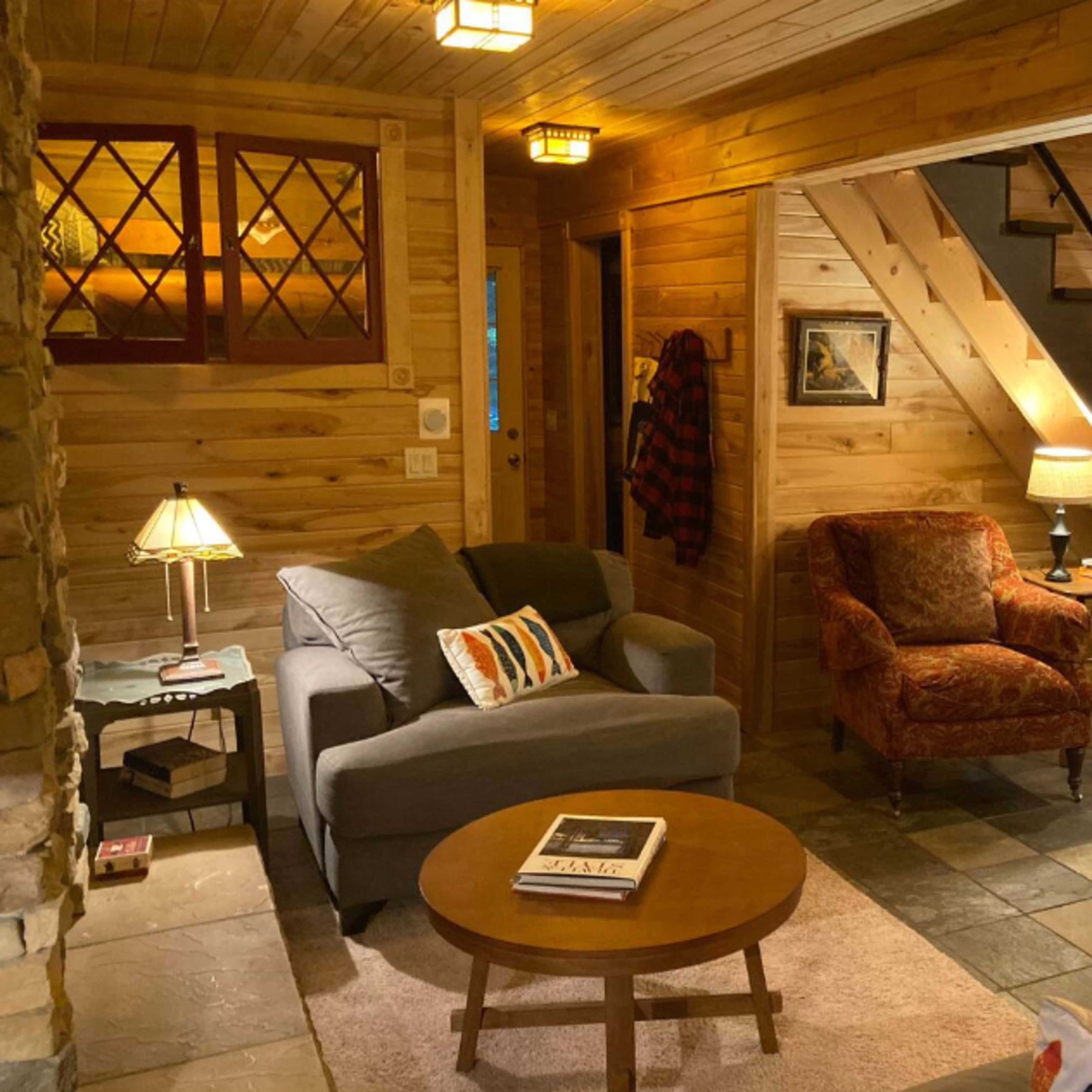 The interior of a wooden-paneled living room features a gray sofa, an ornate armchair, a round coffee table, and a staircase leading to the upper floor.