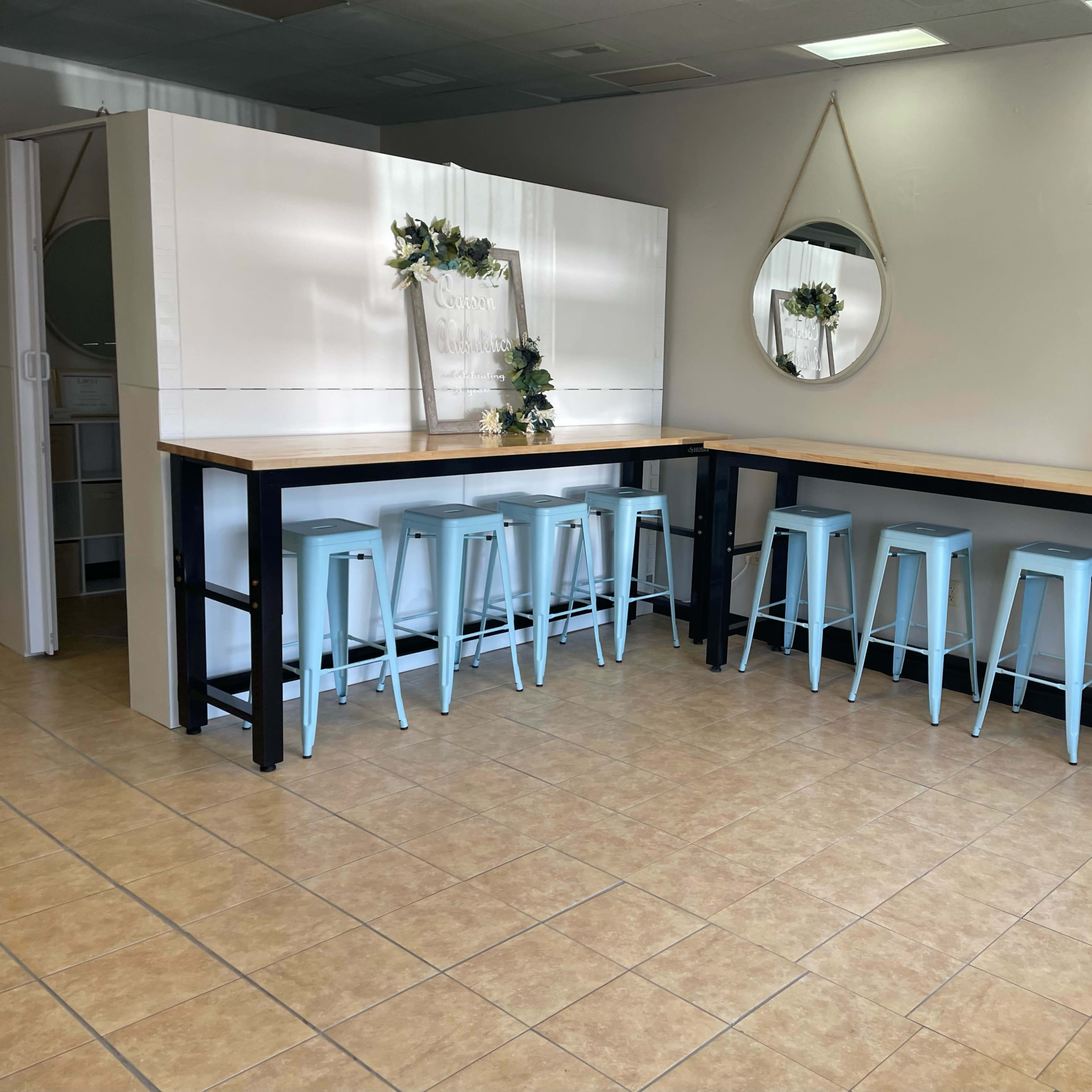 A bright, minimalistic interior featuring two long tables with light blue stools and a round mirror on the wall.