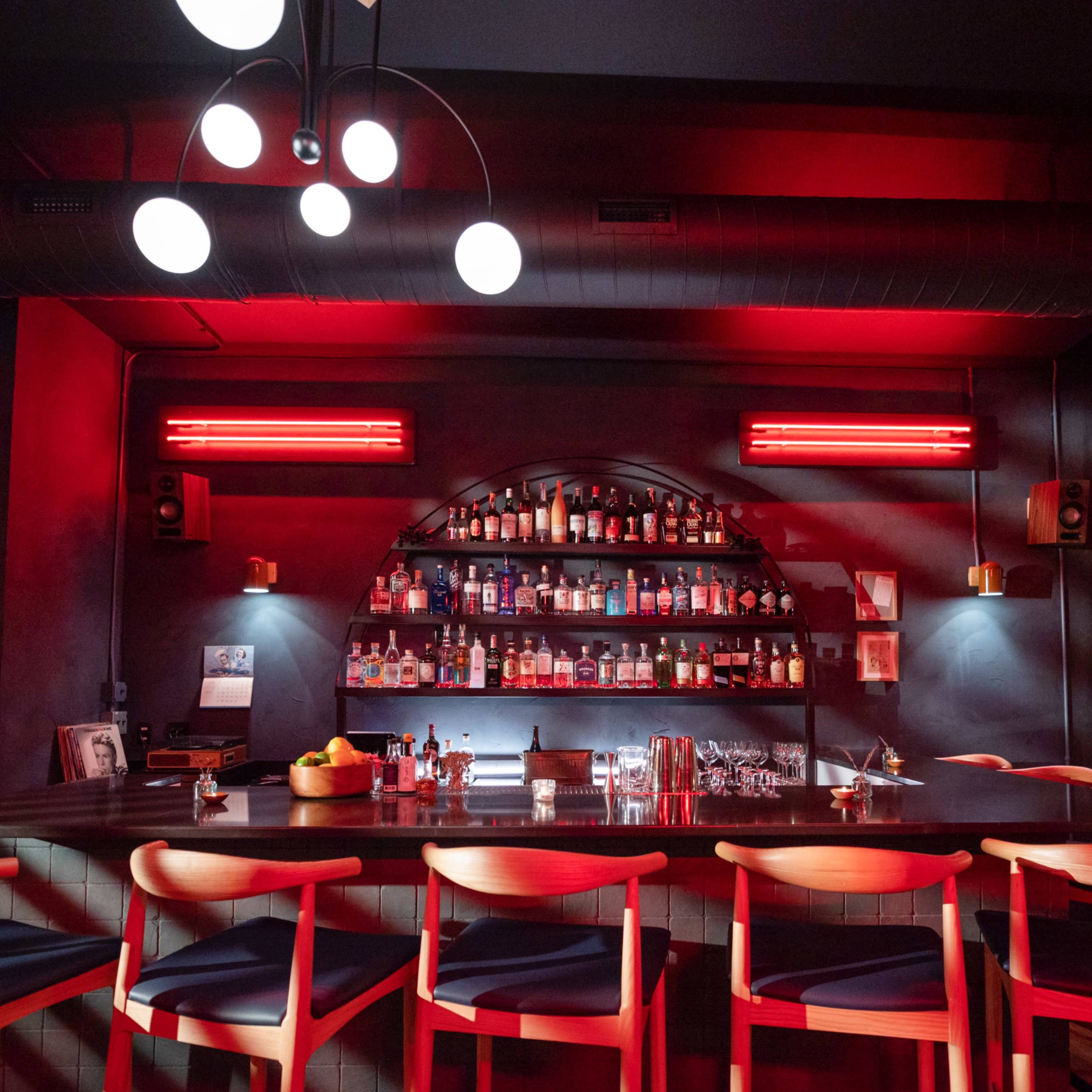A modern bar scene features a long wooden counter with empty chairs, illuminated by red lights, showcasing a wall of various bottles in the background.