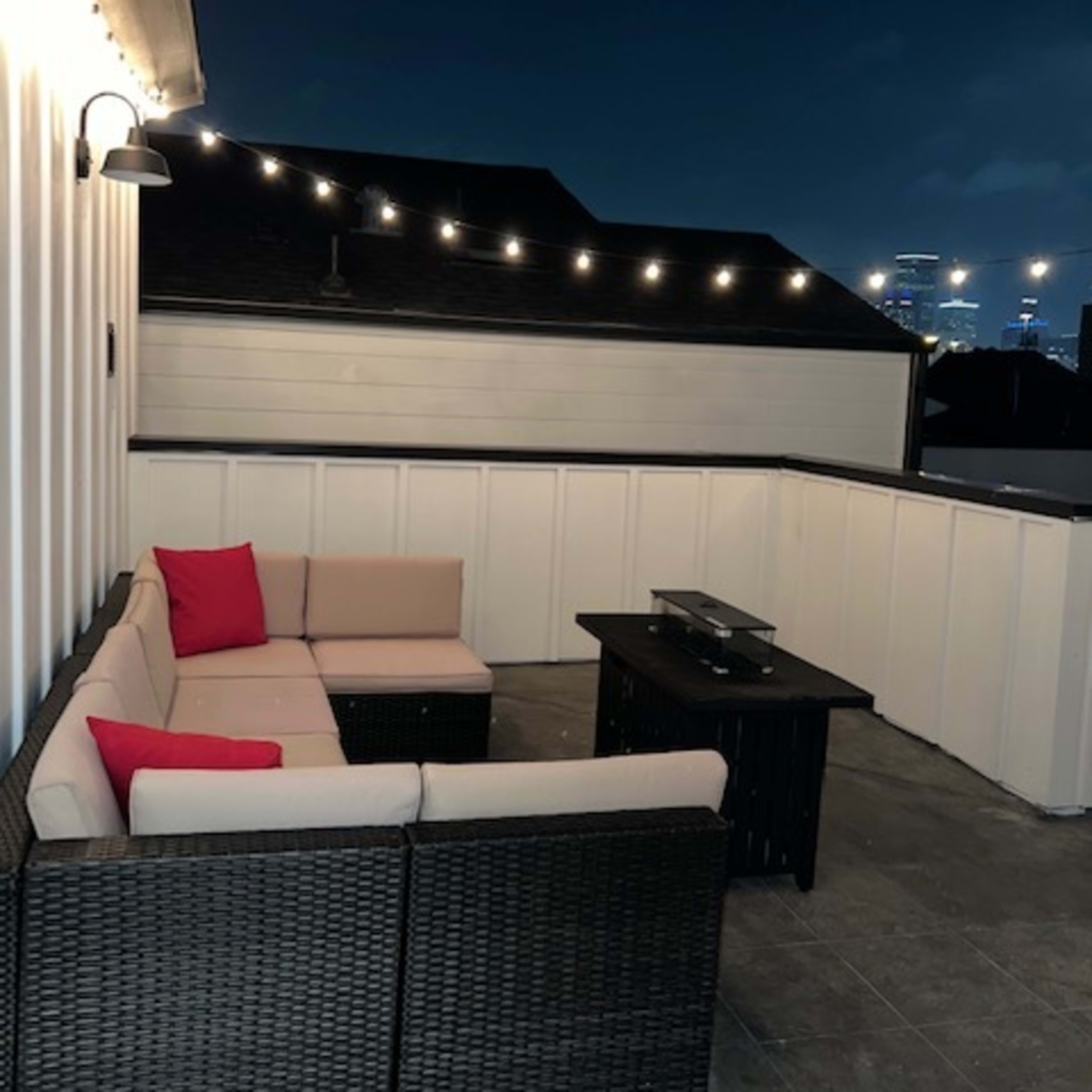 A rooftop seating area features a sectional couch with red accent pillows and a small coffee table, illuminated by string lights against a night sky.