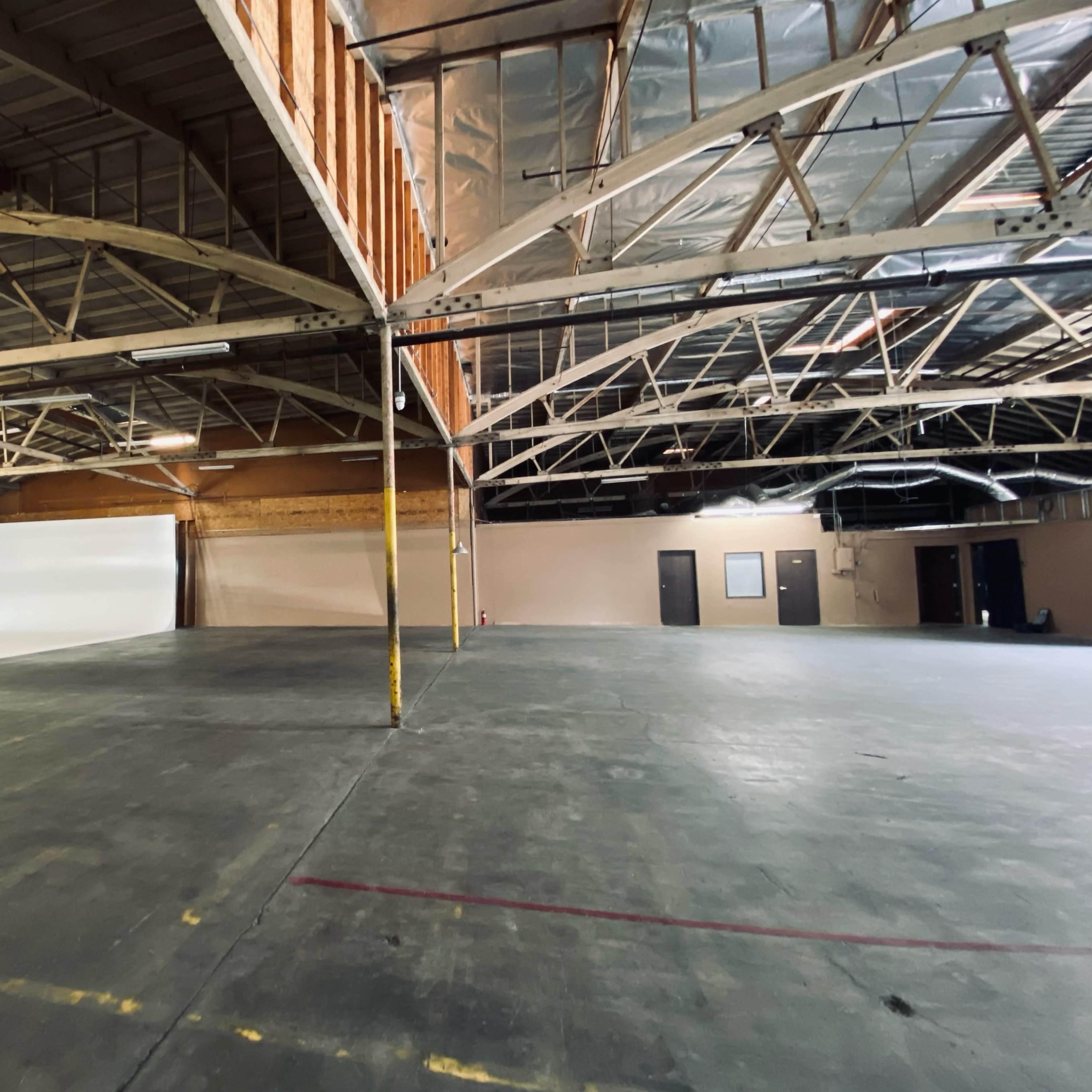 An empty industrial warehouse space with high ceilings and a large, unobstructed area featuring a white backdrop along one wall.
