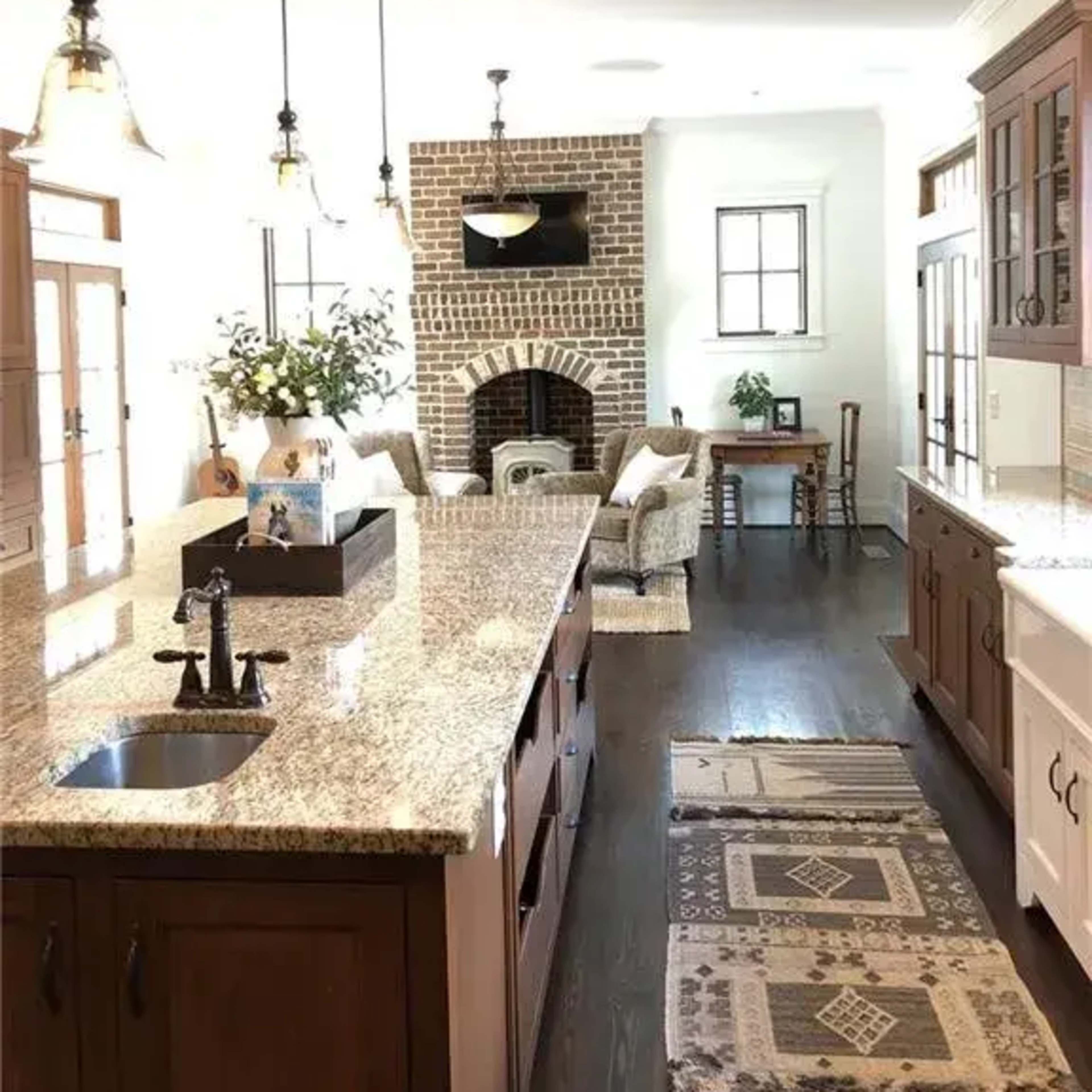 A spacious kitchen features a central island with a granite countertop, wooden cabinetry, and a visible living area with a brick fireplace in the background.
