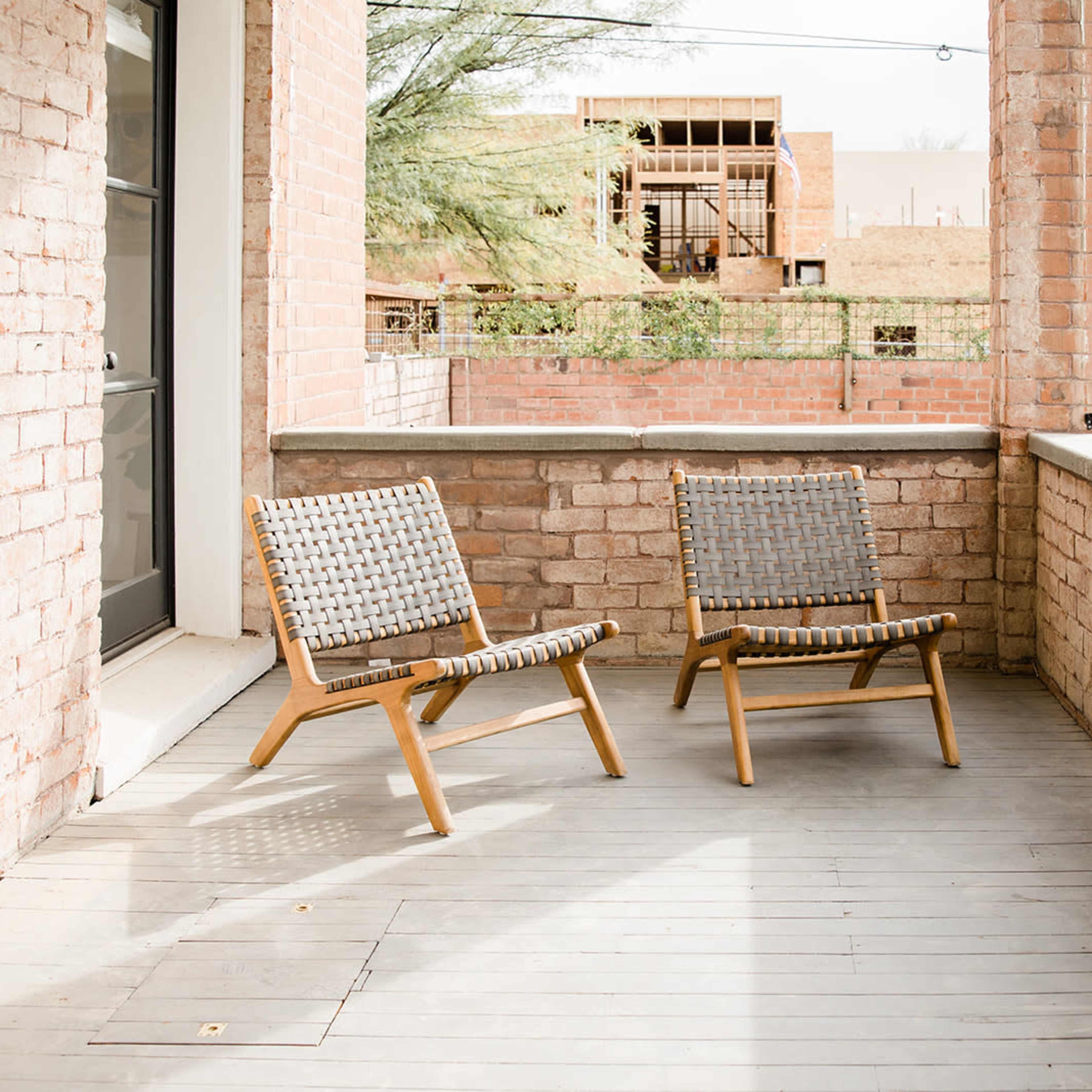 Two wooden-framed chairs with woven seats are positioned on a sunlit balcony surrounded by brick walls.